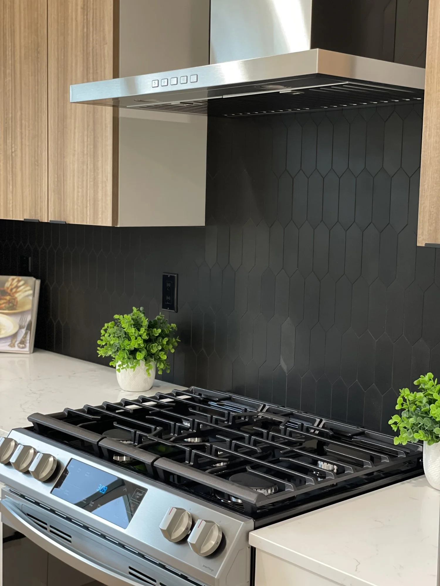 Modern kitchen stove with black hexagon tile backsplash and wooden cabinetry, decorated with potted green plants.