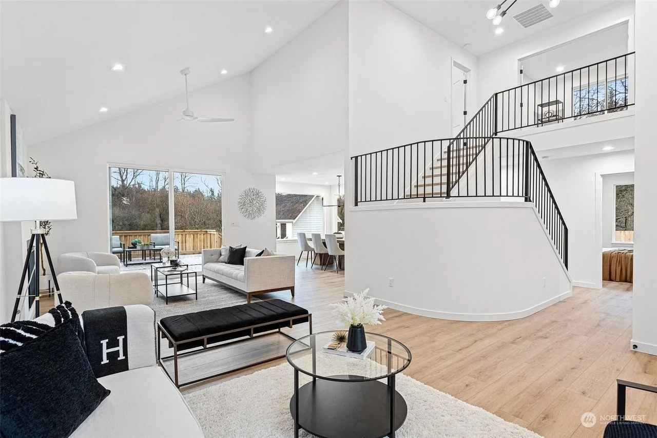 Bright, modern living room with white walls and high ceilings, featuring large windows overlooking a fenced backyard, and furnished with white sofas, a black bench, a round glass coffee table with flowers, and a standing lamp.