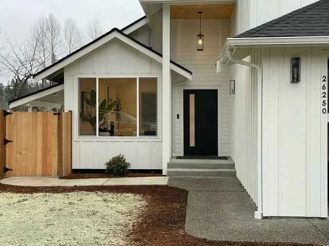 Front view of a modern white house with a black front door, concrete steps, a porch light, a large window, and a wooden fence on the left side.