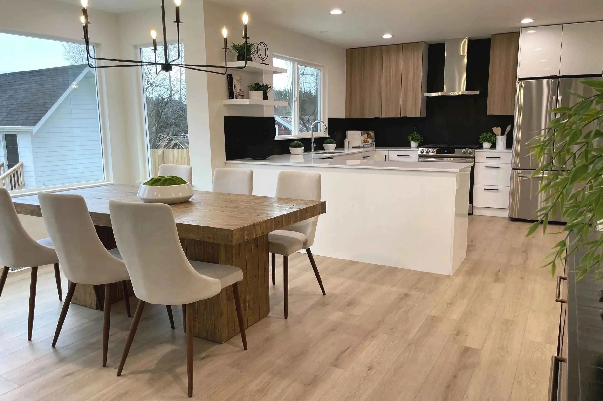 Modern kitchen with white and wood cabinets, stainless steel appliances, a large window, a wooden dining table with six white upholstered chairs, a black chandelier above the table, and decorative plants on the counters and shelves.