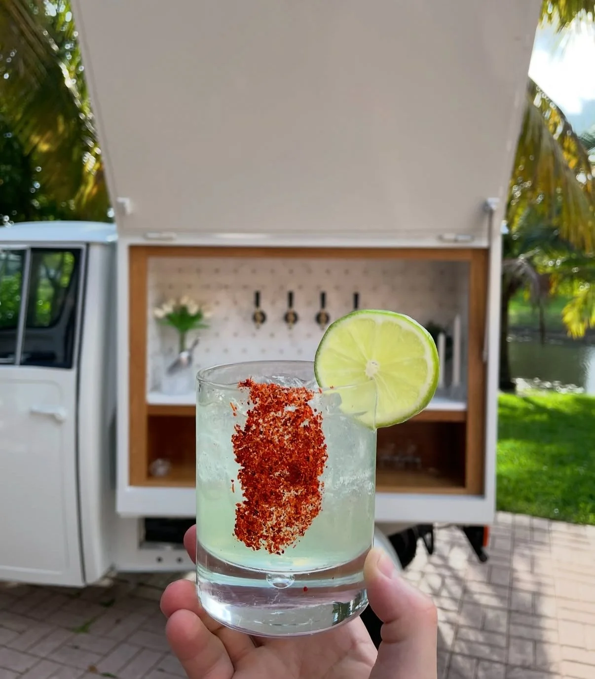Hand holding a lime-garnished craft cocktail in front of Tap Socialās Piaggio Ape mobile bar with a 4-tap self-serve system at a South Florida event