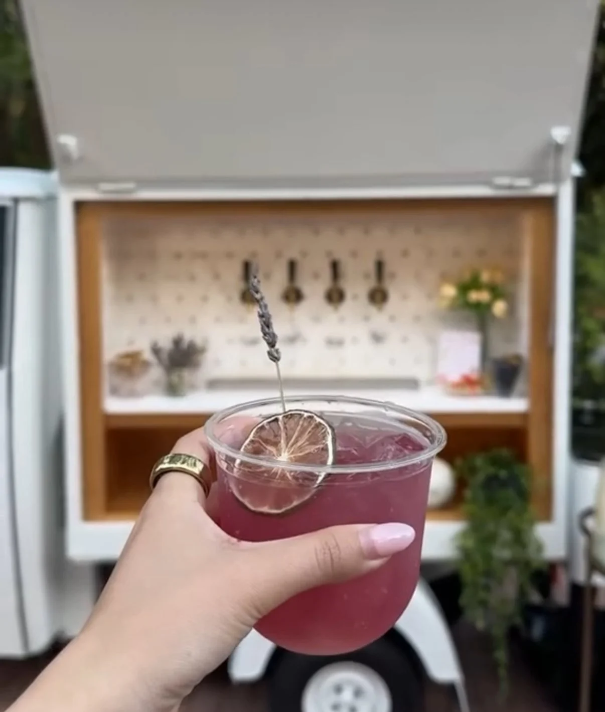 Hand holding a craft cocktail in front of Tap Socialās Piaggio Ape mobile bar with a 4-tap self-serve beverage system at an outdoor event in South Florida.