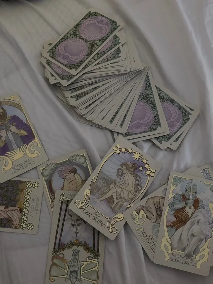 A spread of Tarot or oracle cards on a white cloth, featuring illustrations of a character in armor, a woman with long hair sitting on rocks, and a woman with stars around her head, along with a pile of banknotes.