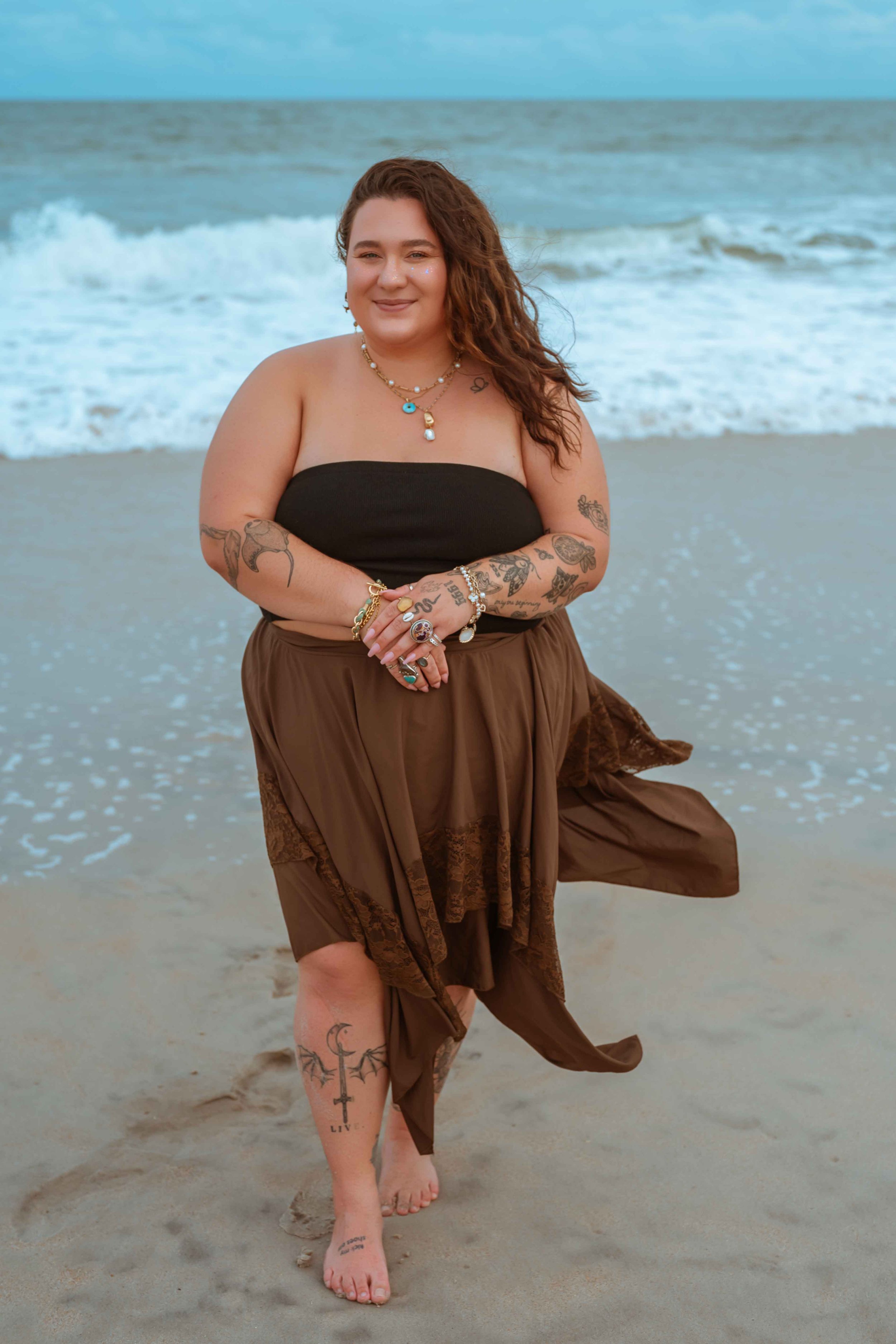 A woman with tattoos on her arms and legs stands on a sandy beach, wearing a black strapless top and a brown skirt, smiling at the camera. The ocean waves are in the background.