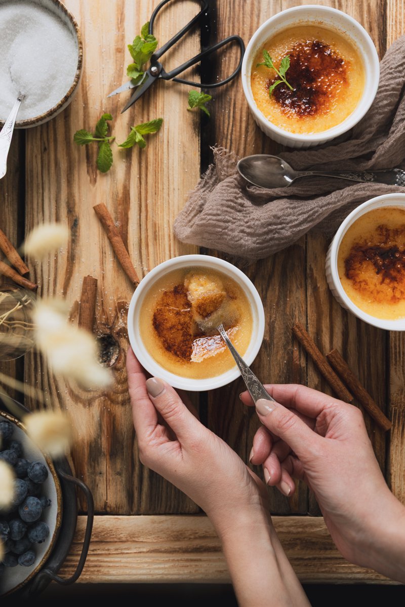 Three baked crème brûlées on a wooden table, one being prepared with a spoon, alongside a sugar bowl, cinnamon sticks, fresh blueberries, and some mint leaves.