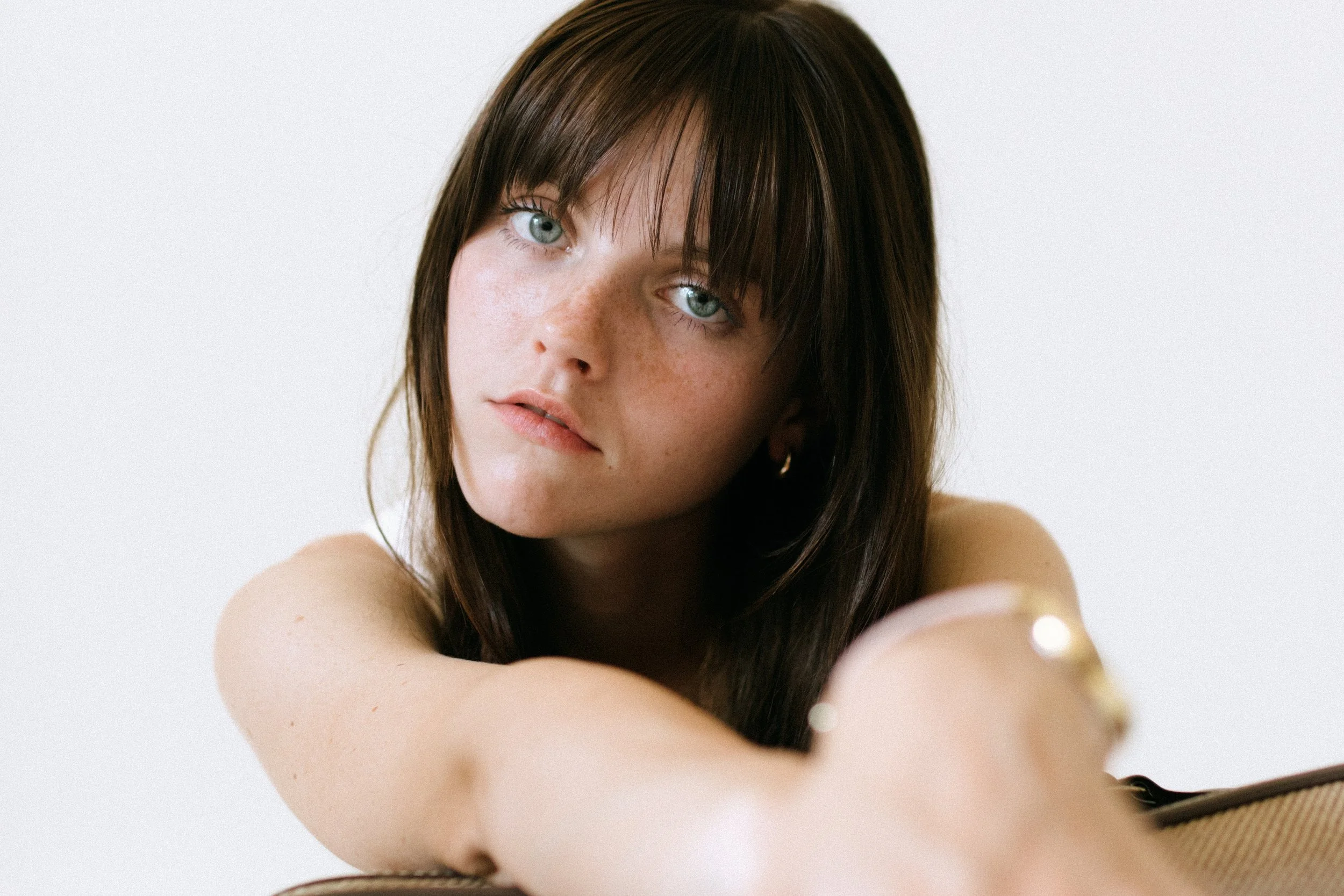 Young woman with long brown hair and bangs, wearing gold hoop earrings, looking directly at the camera.