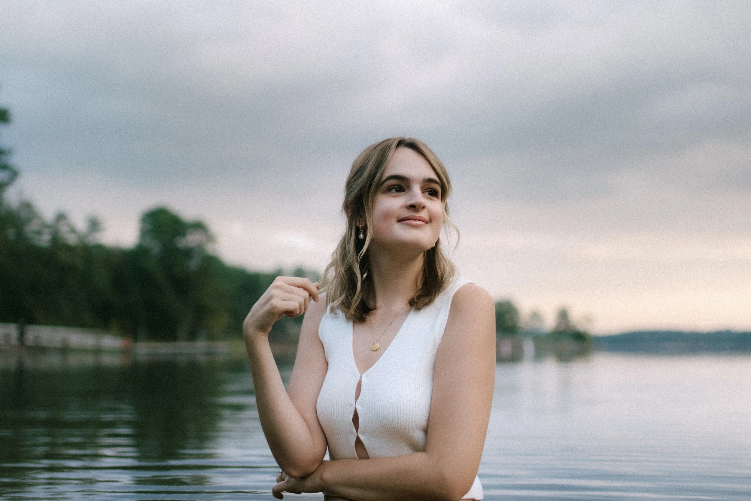 Woman in white top standing by a lake with trees in the background, cloudy sky.