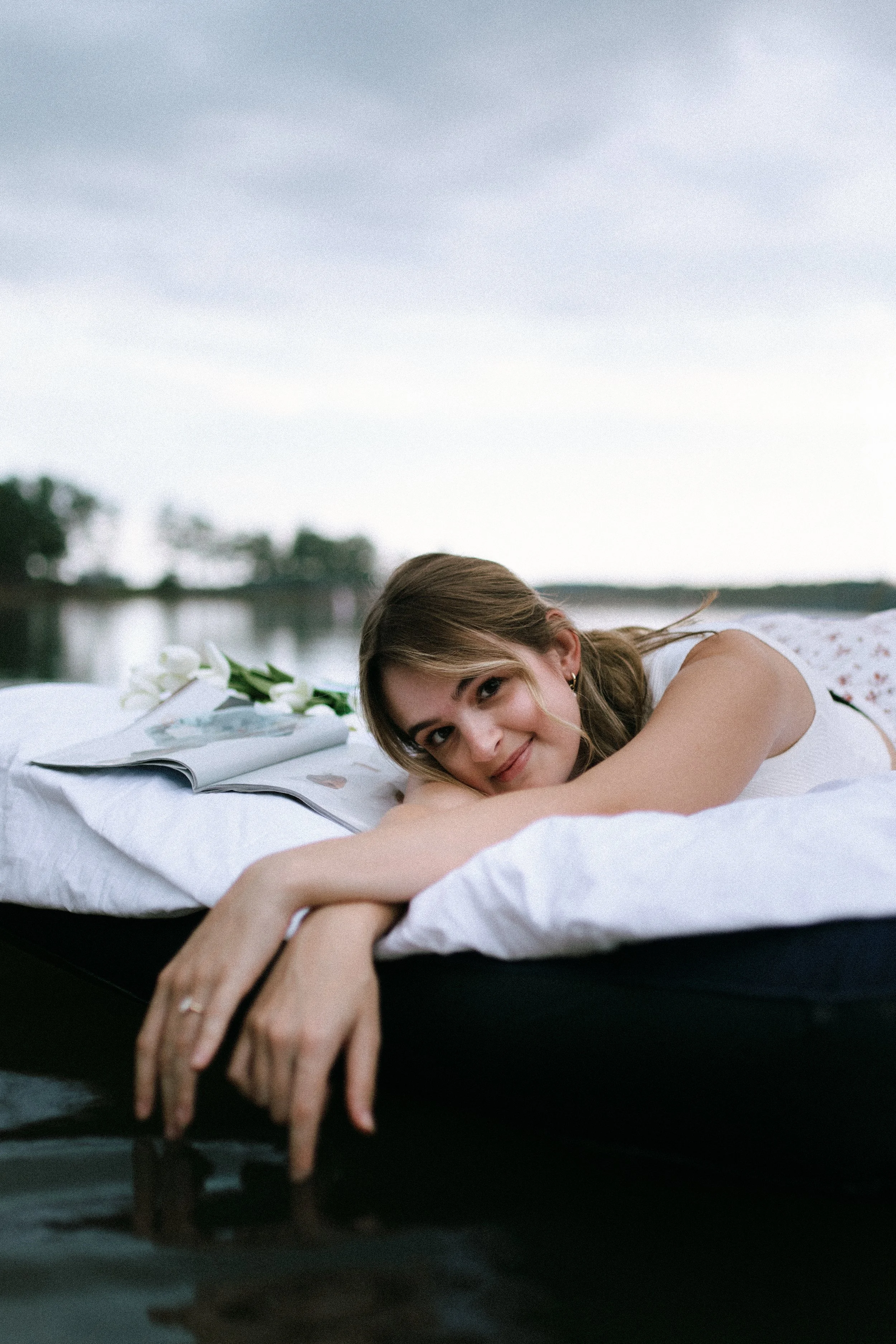 Young woman resting on a floating bed on a lake, with an open magazine and lilies beside her, smiling with her hand touching the water.