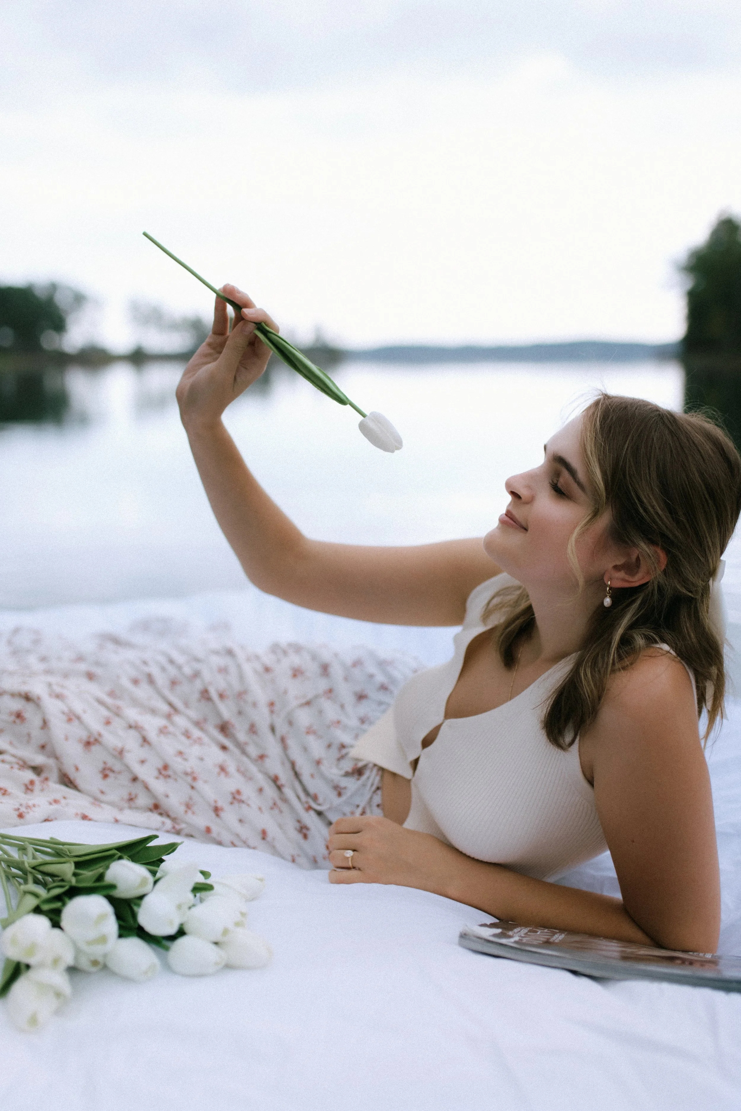 Woman laying on a blanket by the water, holding a white tulip, with more white tulips in the foreground.