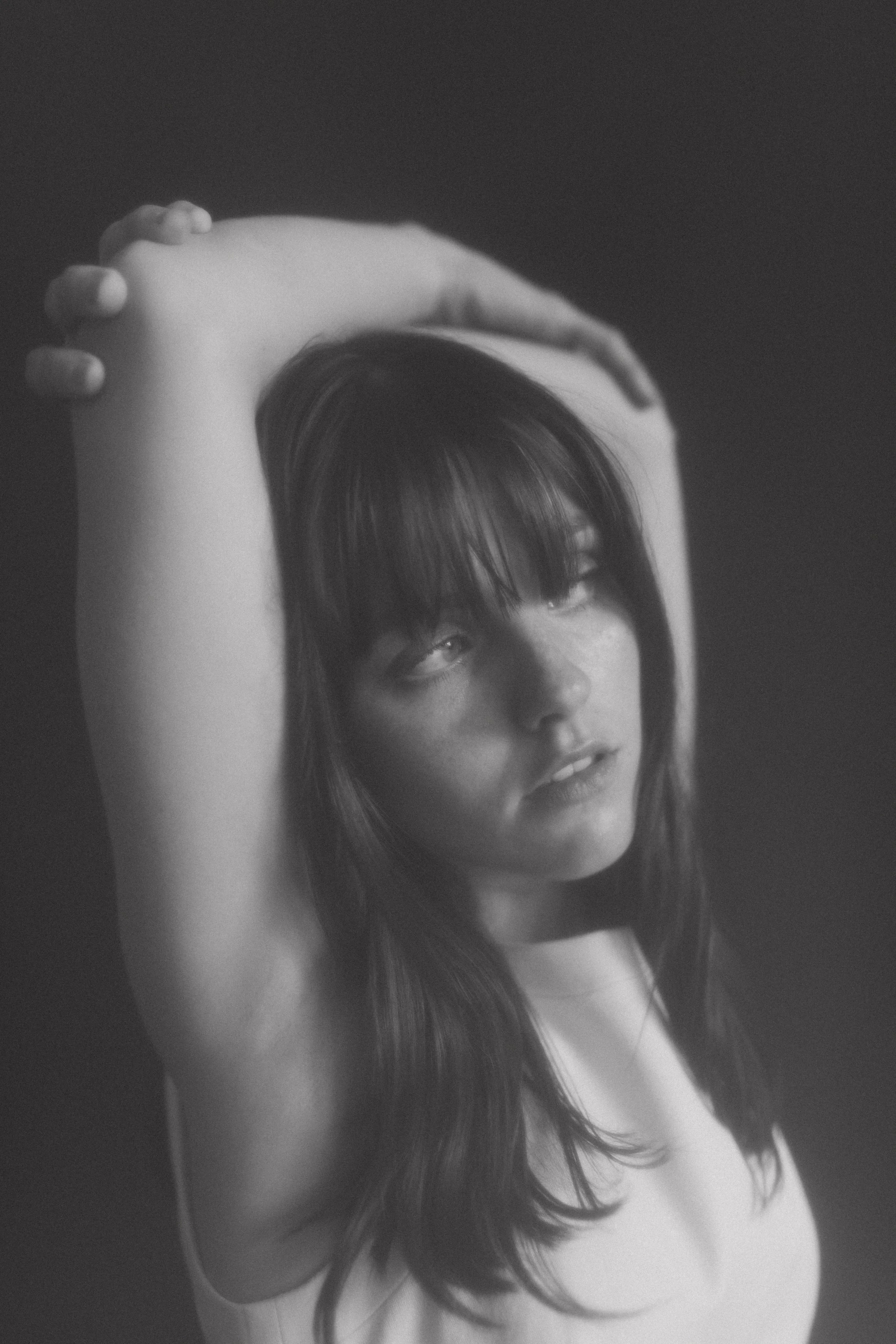 Black and white portrait of a woman with long hair, arms raised, looking to the side.