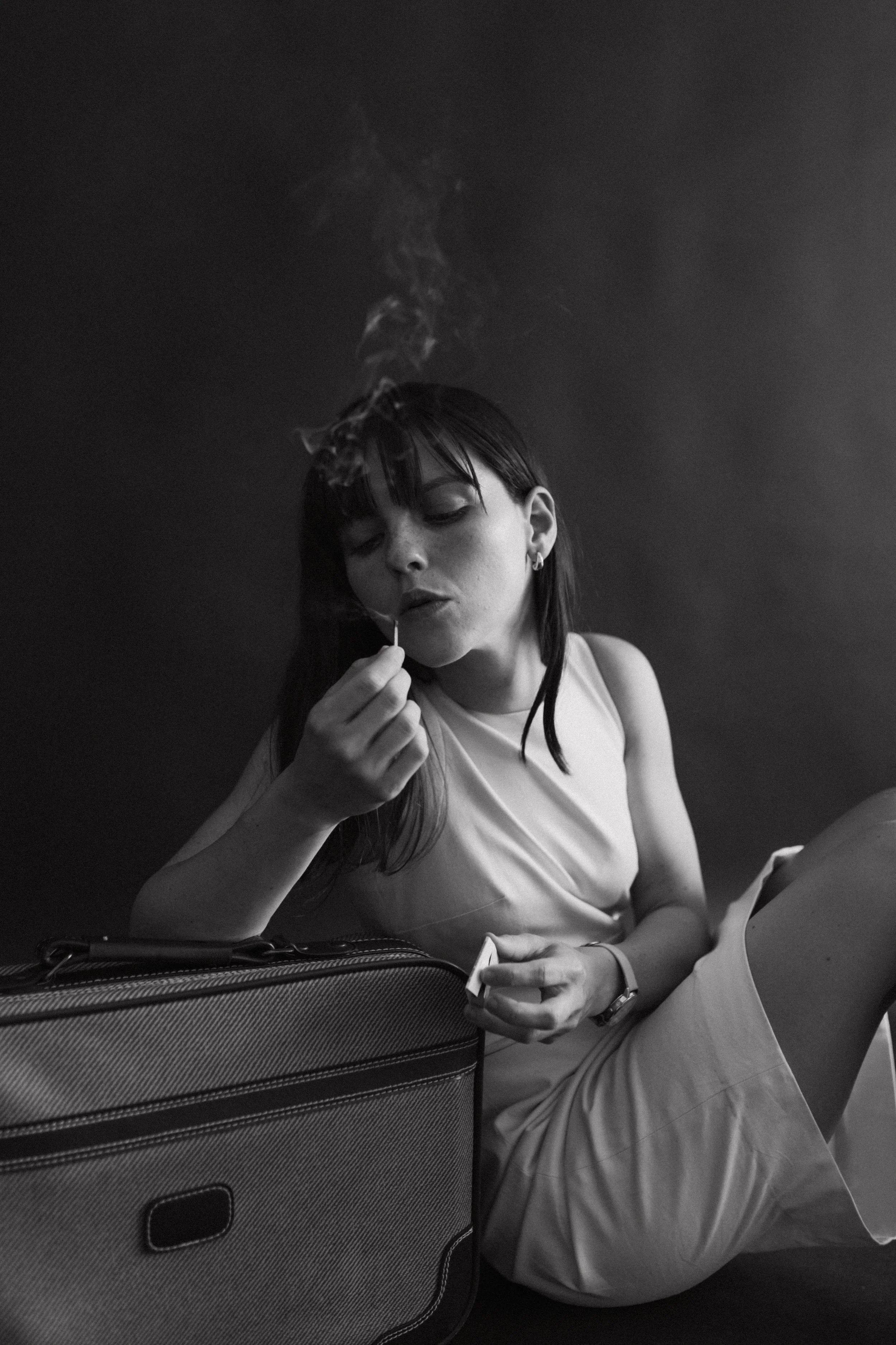 Woman in white dress lighting a cigarette next to a suitcase in black and white