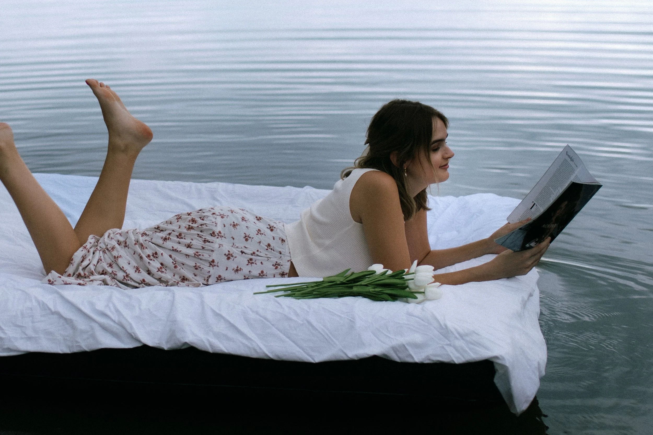 Woman relaxing on floating bed in water, reading a magazine, with a bouquet of white tulips.