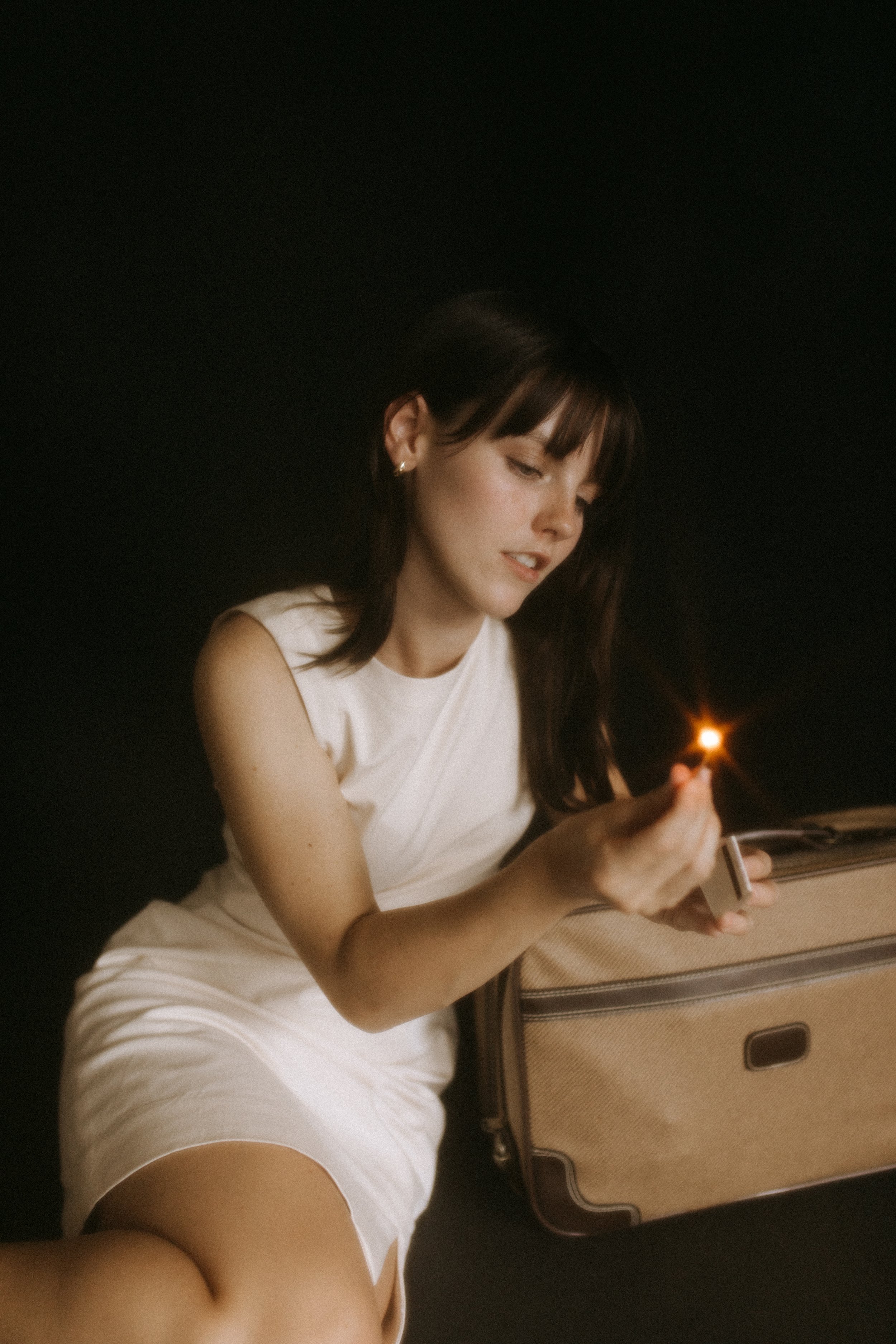 Woman in white dress holding a matchstick near a suitcase in a dimly lit setting.