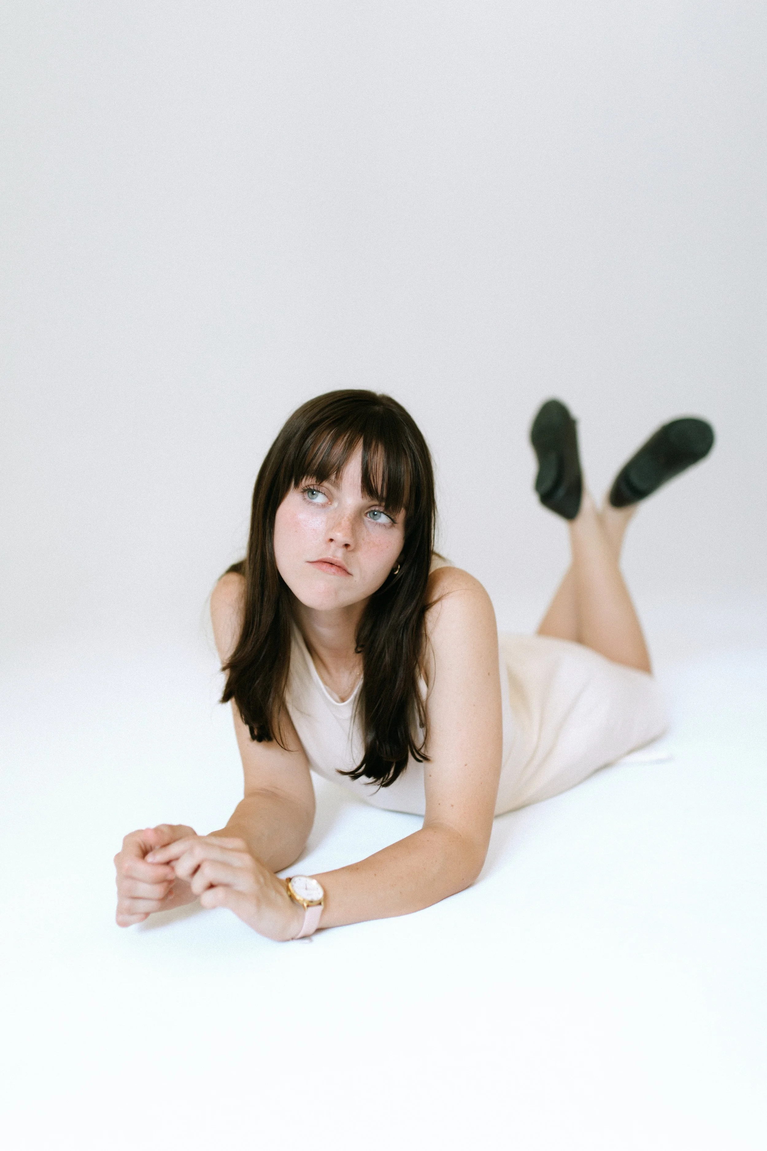 Woman with dark hair lying on the floor, wearing a beige sleeveless dress and black shoes, looking upwards.