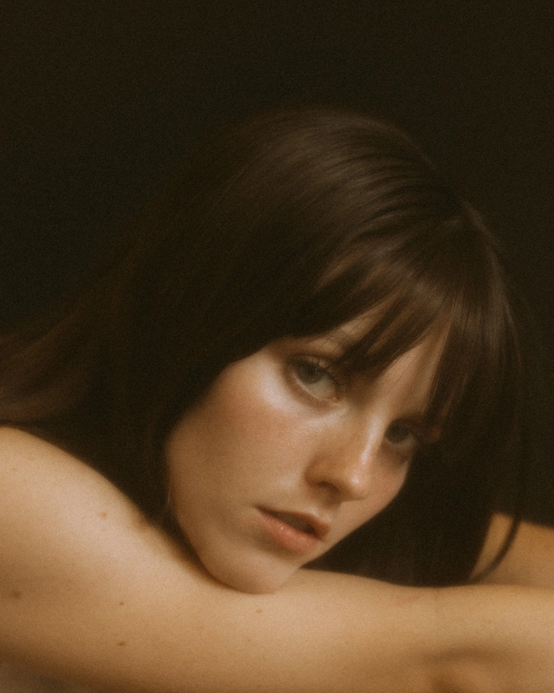 Portrait of a woman with long hair and bangs, gazing towards the camera with a pensive expression, set against a dark background.