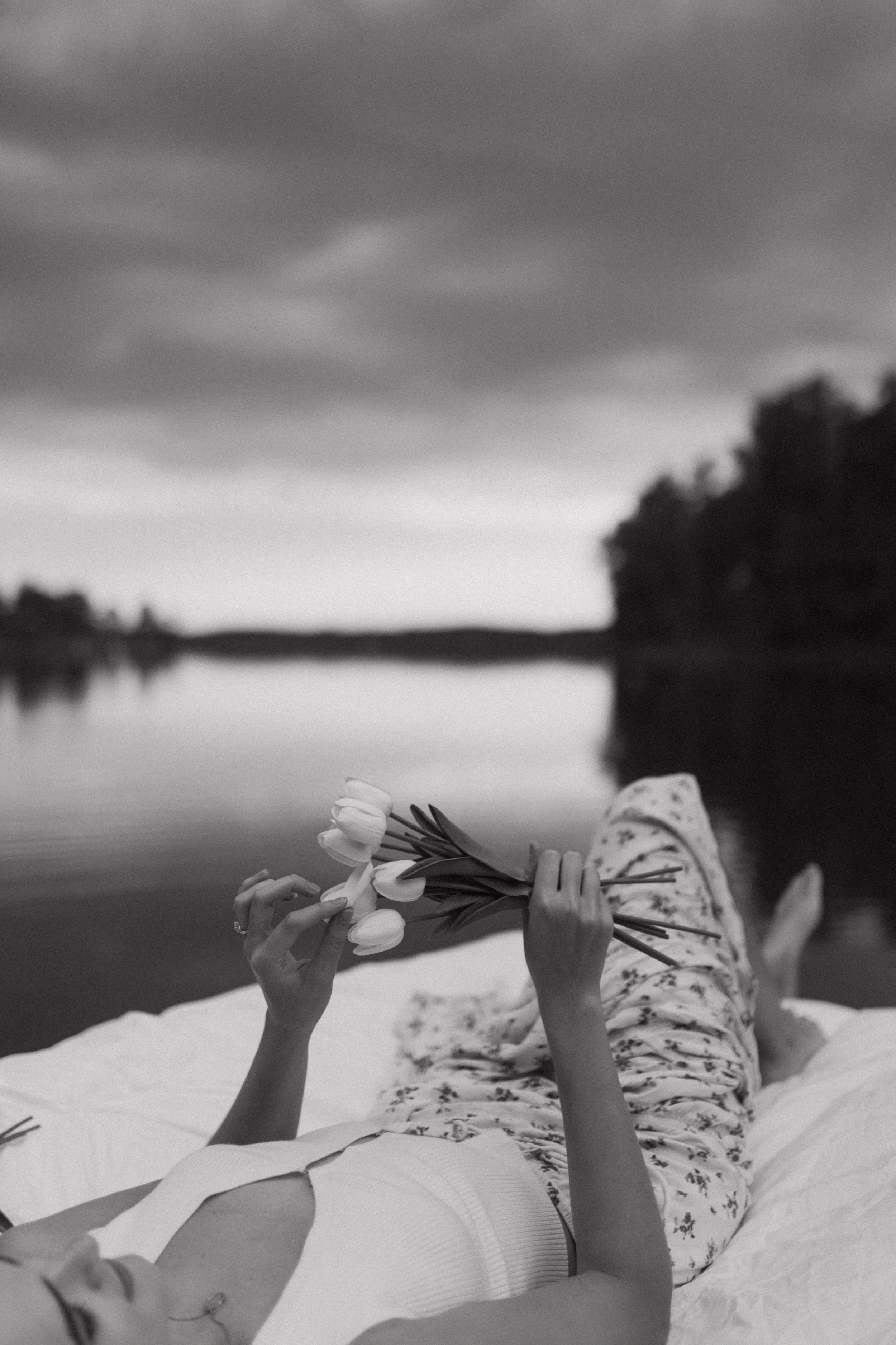 Black and white image of a person lying on a blanket near a lake, holding a bouquet of tulips.