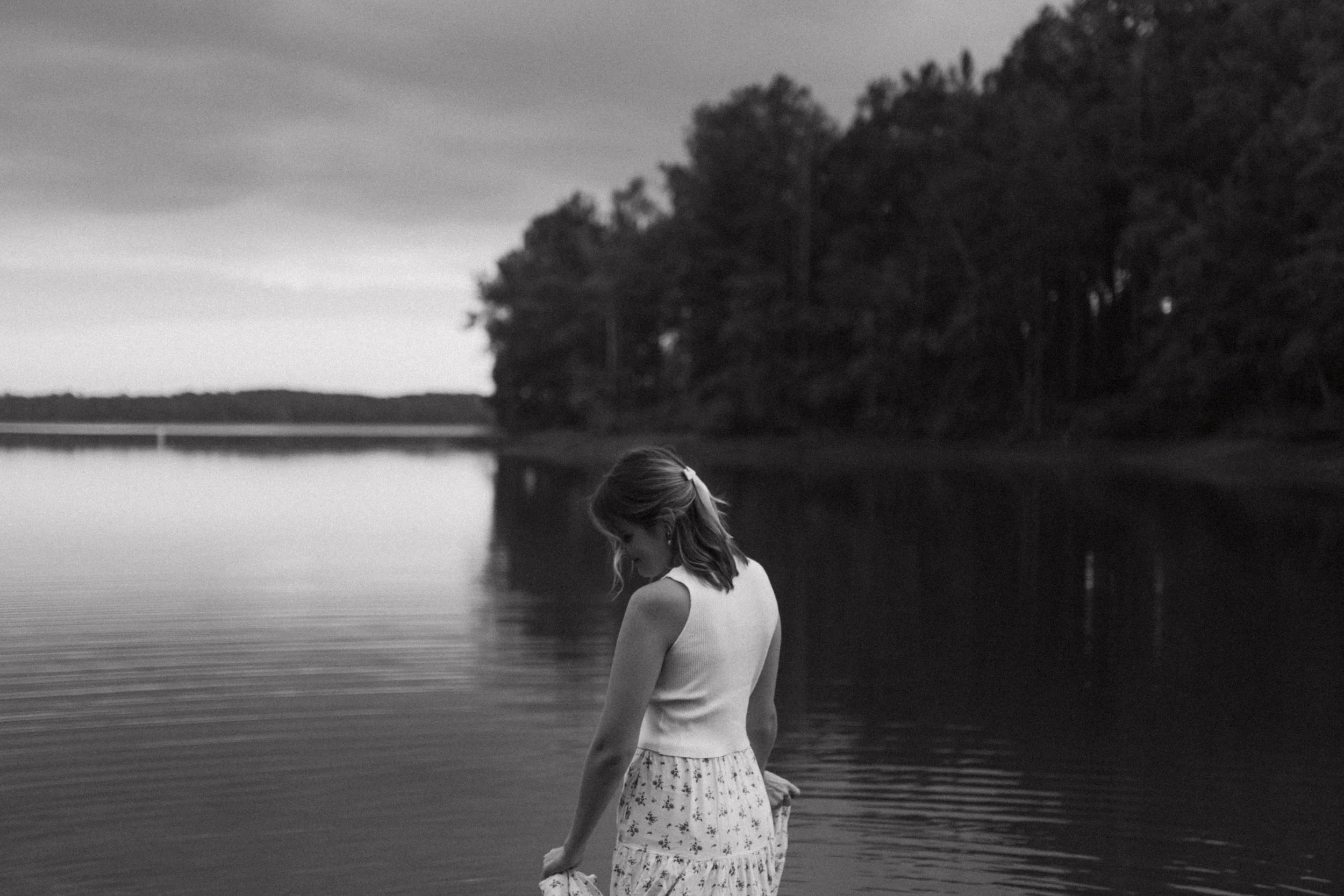 Black and white photo of a woman by a tranquil lake, surrounded by trees.