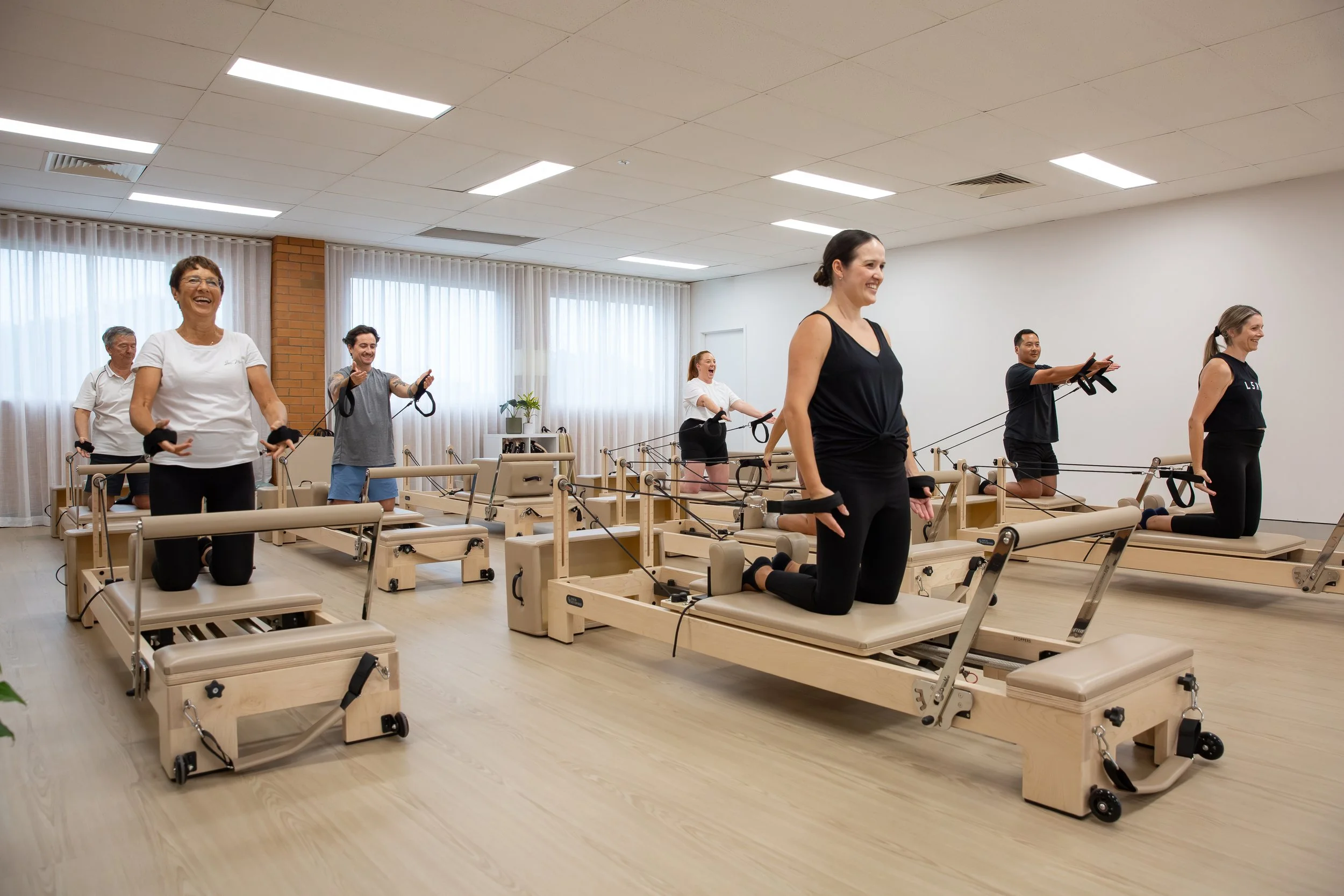 A group of people participating in a Pilates class, kneeling on Pilates reformer machines in a bright, spacious room with white walls and large windows with curtains.