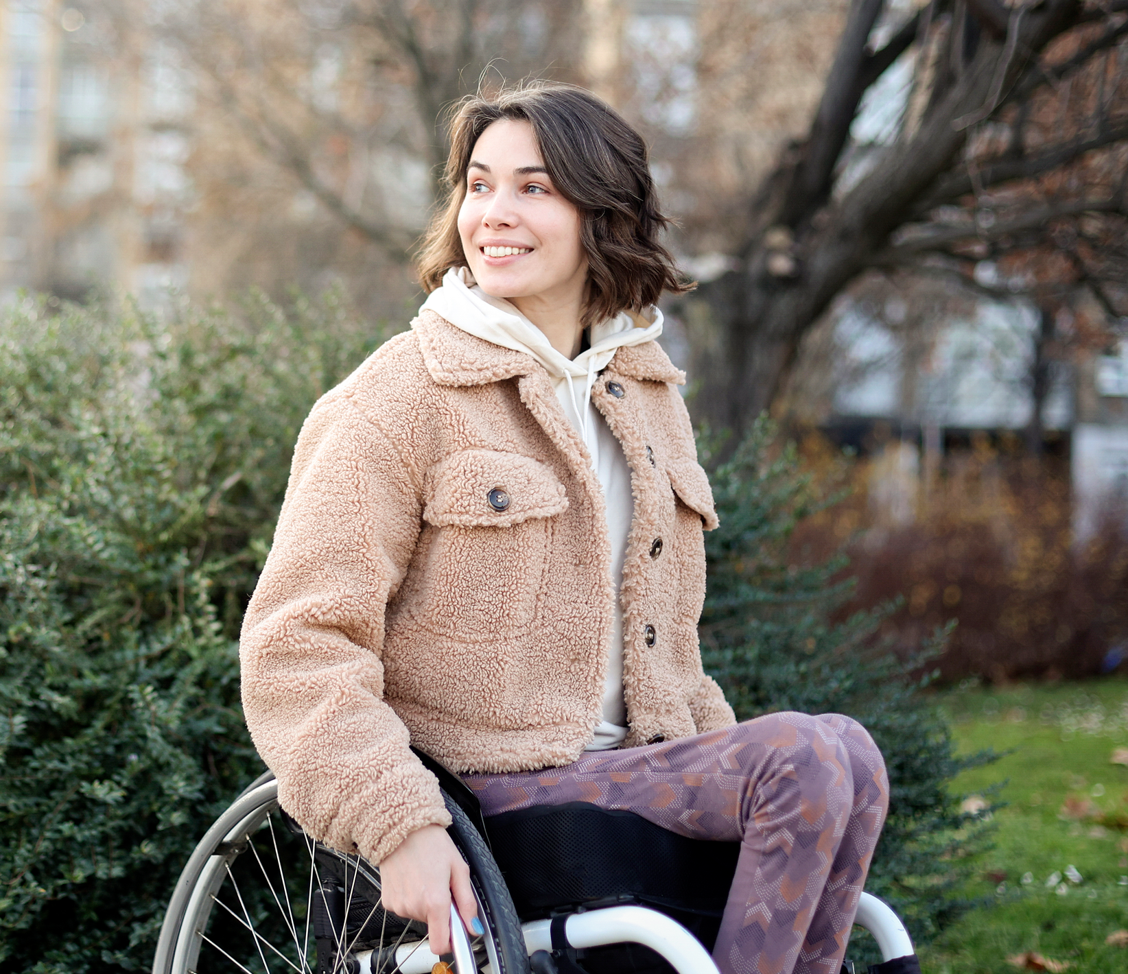 A woman sitting in a wheelchair outdoors in a park, smiling and looking to her left. She is wearing a tan teddy coat, patterned pants, and a hoodie.