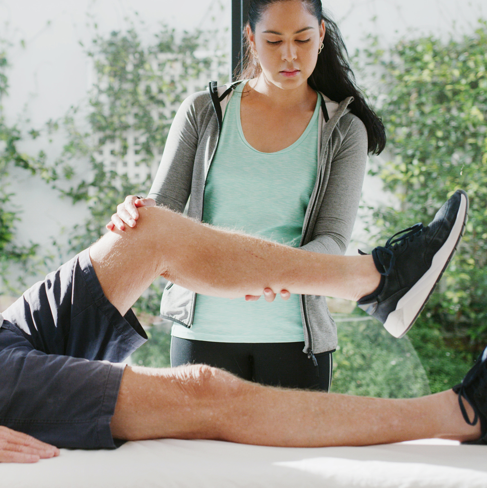 A woman is performing physical therapy on a man's leg, which is extended on a table, in a bright outdoor setting.