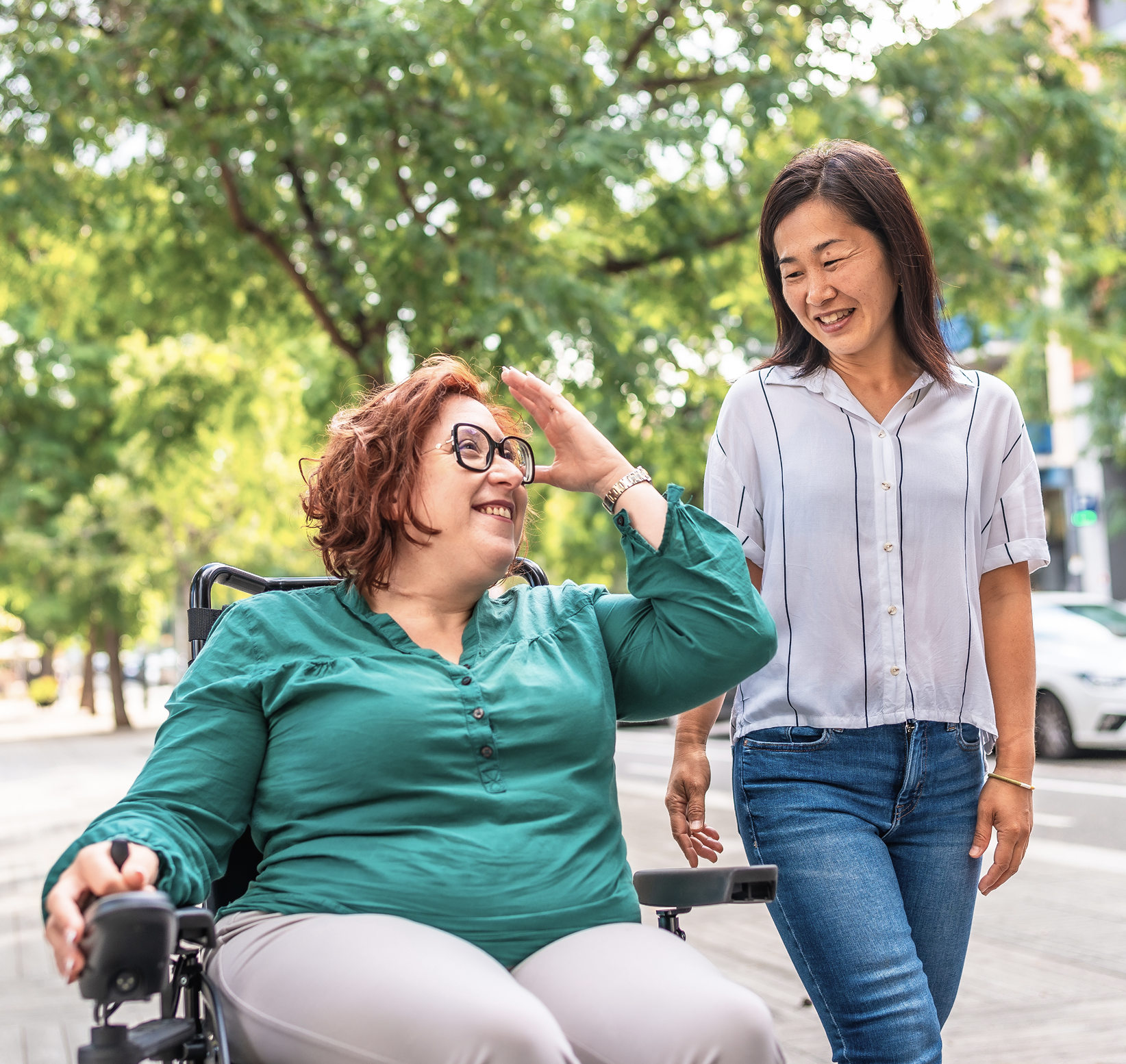 Two women, one in a wheelchair and the other standing, smiling and sharing a moment outdoors under green trees.