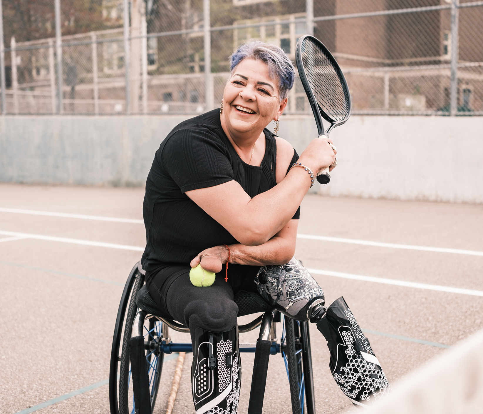 A woman with short gray hair in a wheelchair on an outdoor tennis court, holding a tennis racket and smiling.
