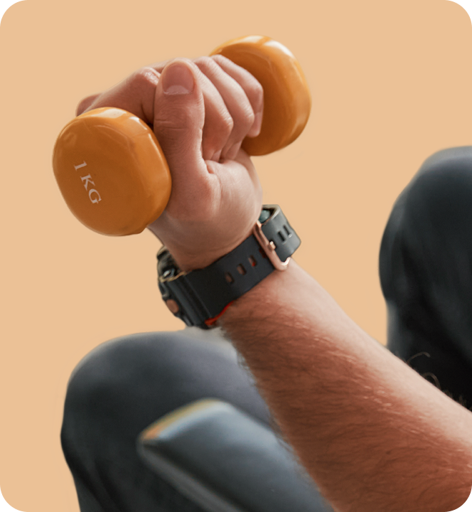 Close-up of a person's hand gripping an orange 1-kilogram dumbbell during exercise, with a watch on their wrist and part of a chair or bench in the background.