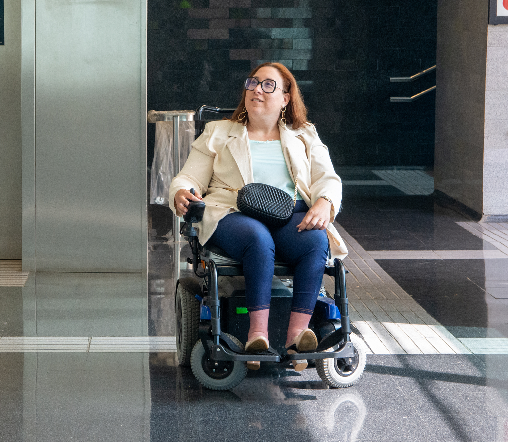 Woman in a wheelchair sitting in an indoor public space, looking up and smiling.