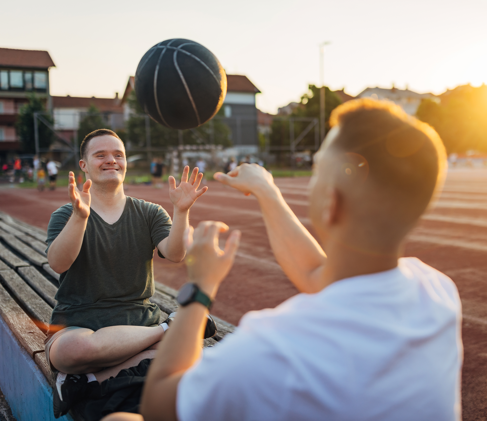 Two young men playing with a basketball on a court during sunset, sitting on a wooden bench, with houses and trees in the background.
