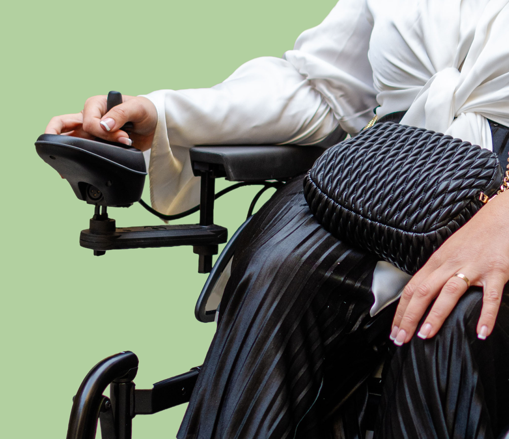 Close-up of a person sitting in a wheelchair, wearing a white blouse and black pleated skirt, with a black woven purse on their lap, holding the wheelchair's control device.