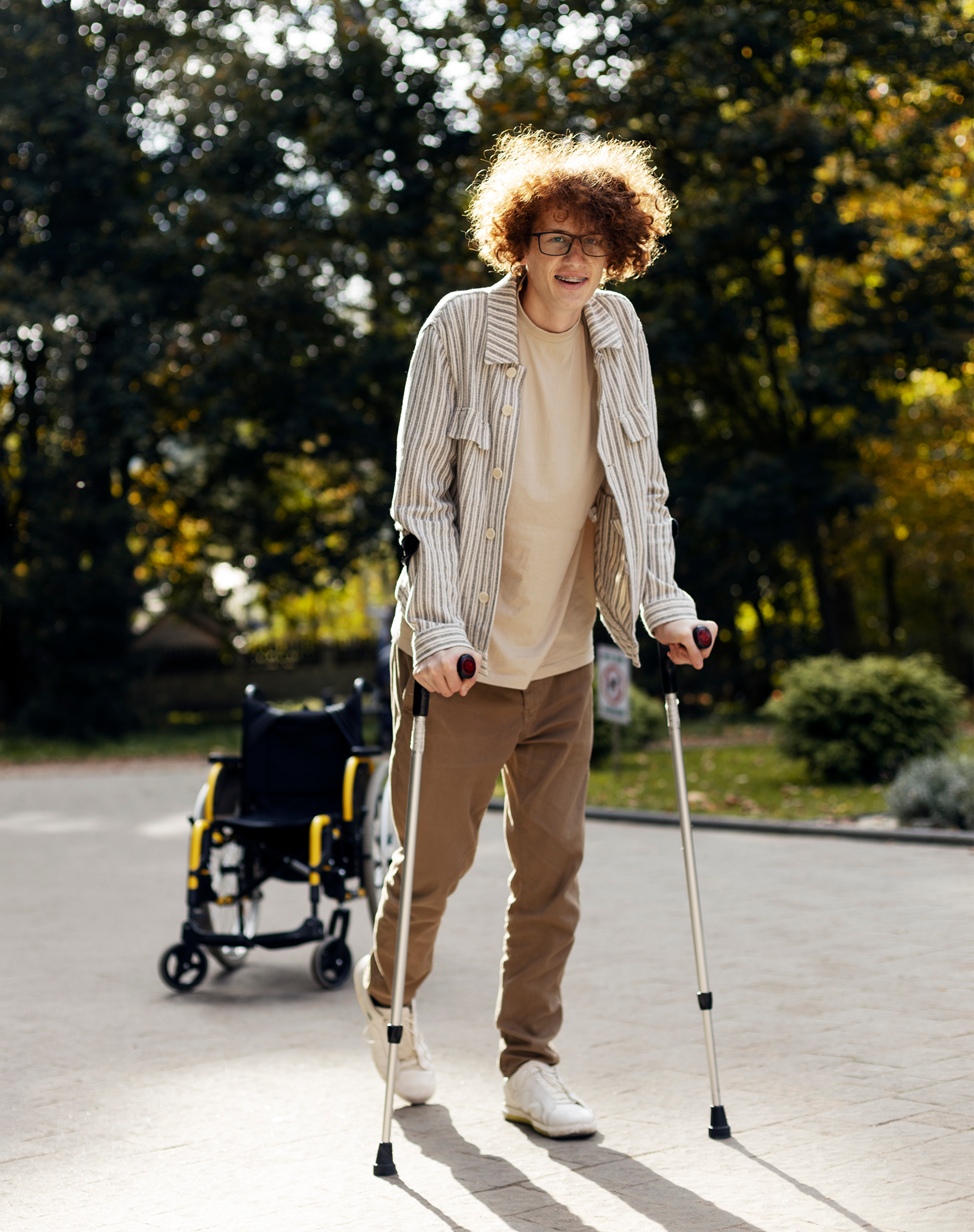A young person with curly hair, glasses, and a striped shirt is using crutches while walking outdoors, with a wheelchair in the background, under trees with autumn foliage.