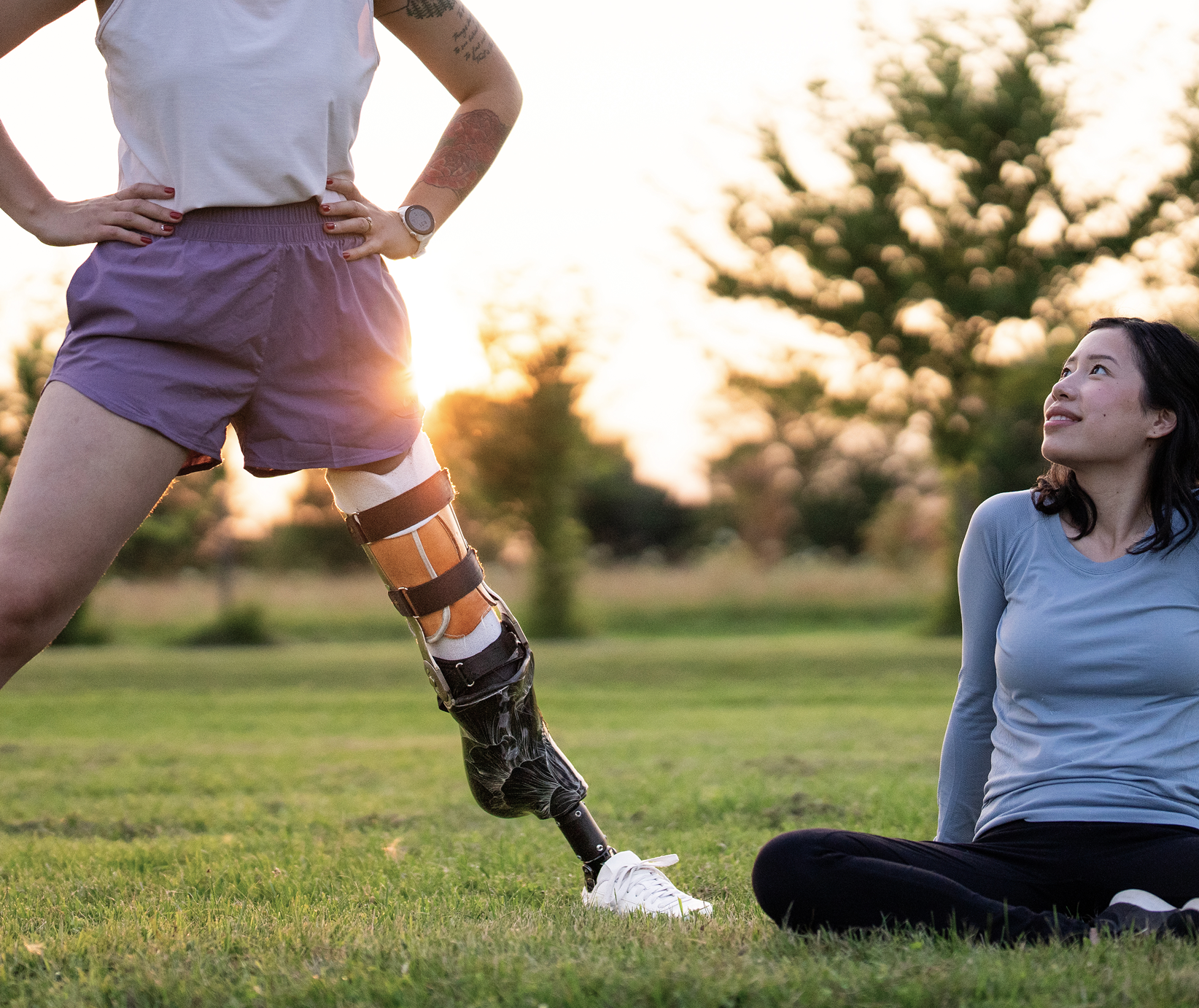 A woman with a prosthetic leg standing outdoors during sunset, with another woman sitting on the grass looking up at her, surrounded by trees.