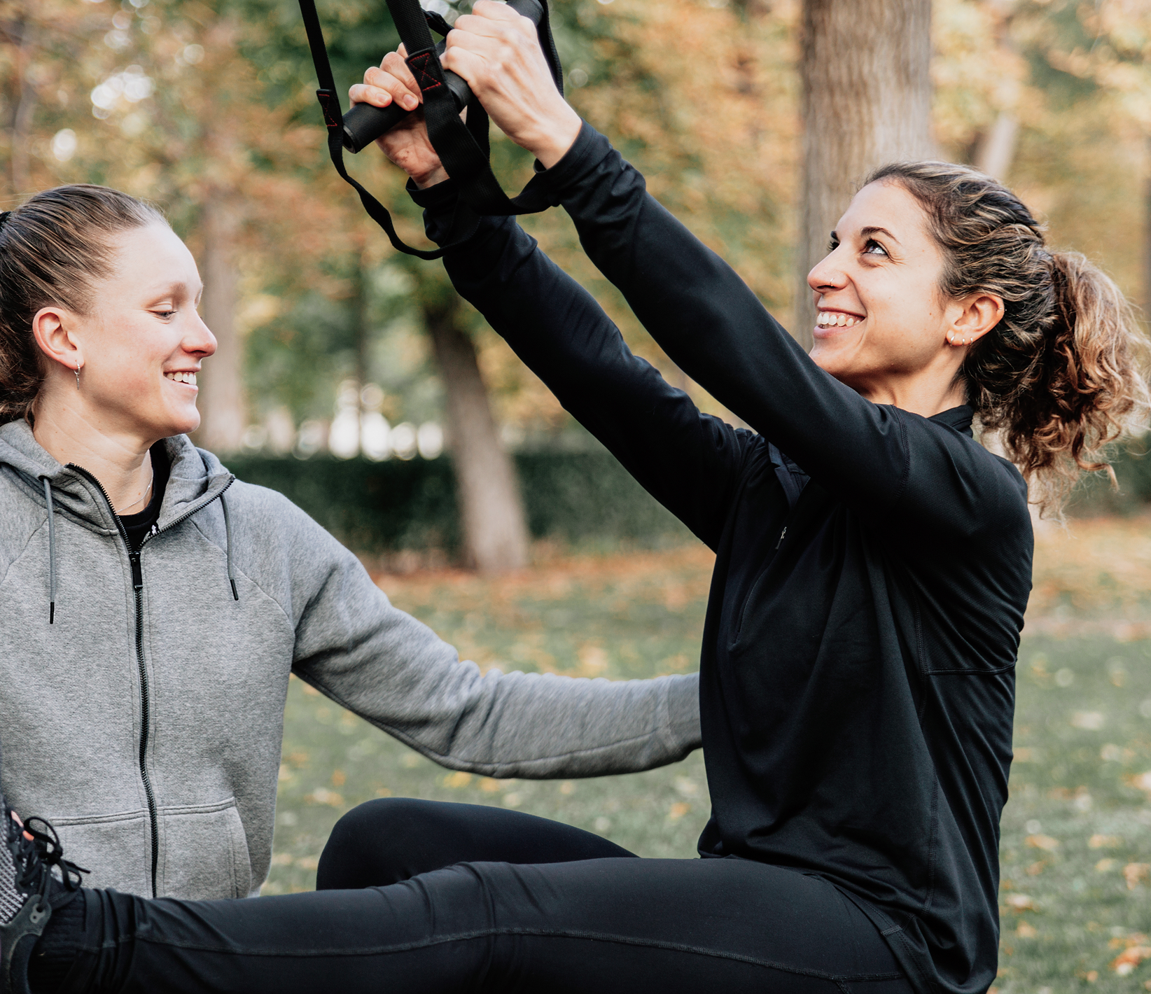 Two women outdoors in a park during autumn, one woman is sitting on the ground and the other is standing and holding a strap above her head. Both women are smiling and wearing casual sportswear.