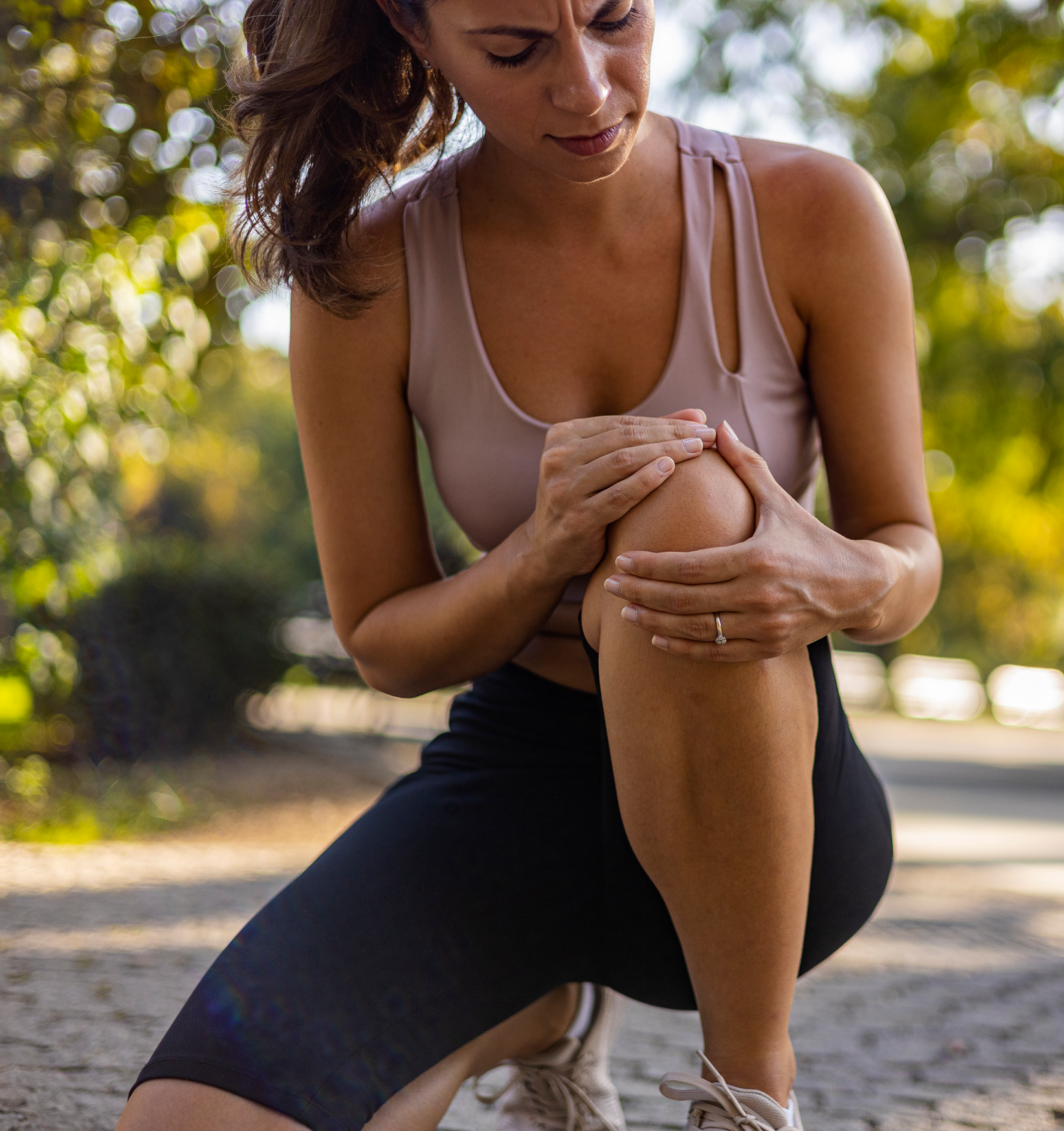 A woman in workout clothes kneeling outdoors, examining her knee with a concerned expression, in a park-like setting with trees and sunlight in the background.