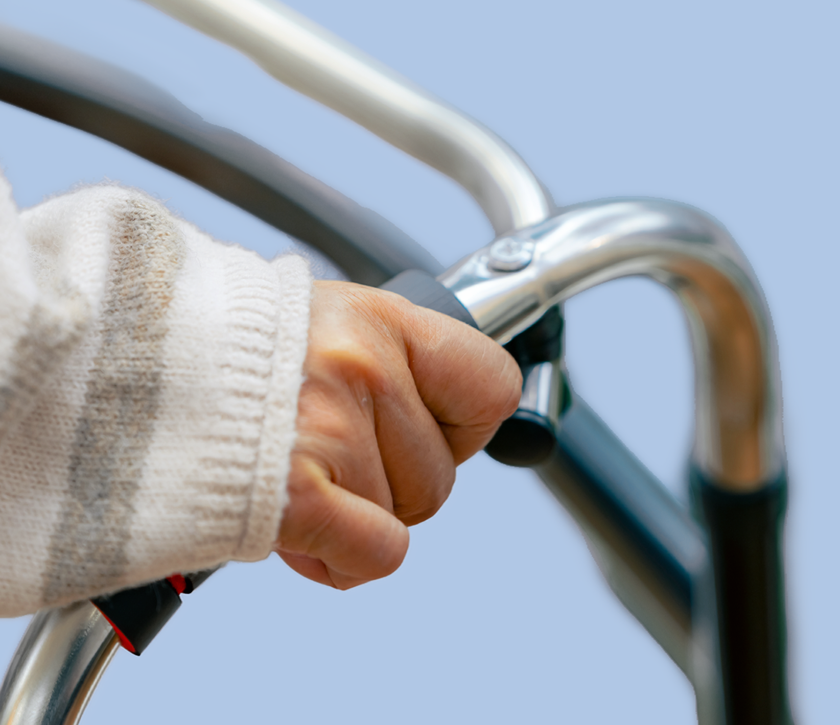 Close-up of an elderly person's hand gripping a walker handle, wearing a white knit sweater, against a light blue background.