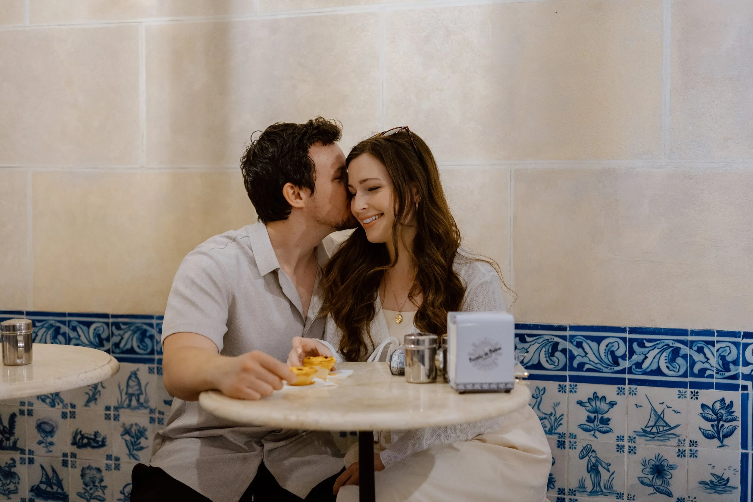 Man kissing his wife’s temple at a café table in front of blue Portuguese tile in Lisbon