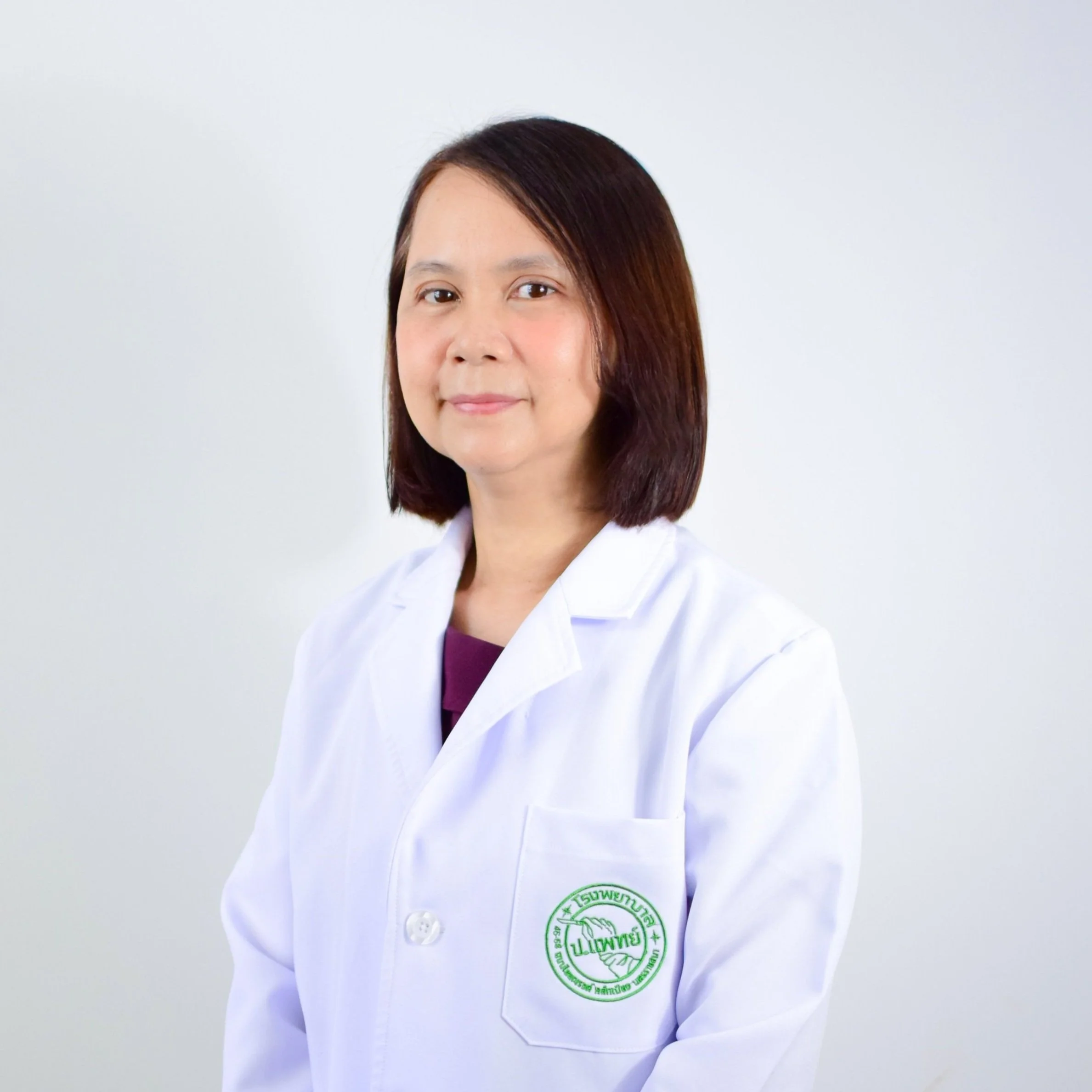 A woman wearing a white medical coat with a green emblem, standing against a plain white background.