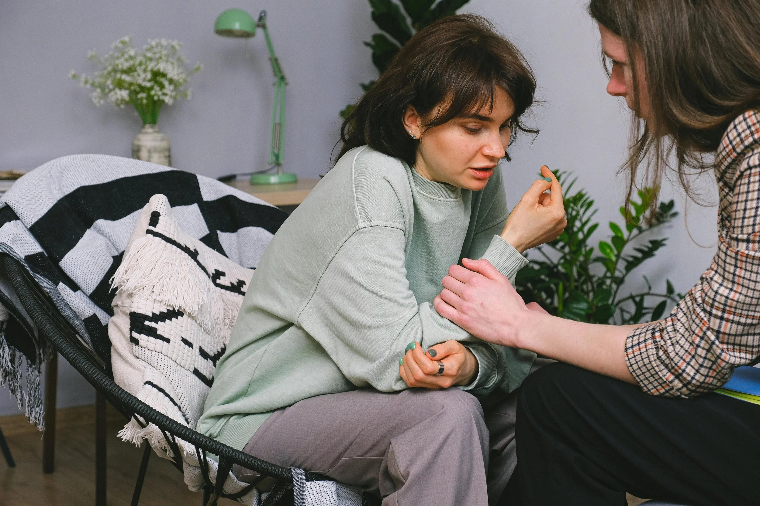 A woman with short dark hair sitting on a chair, holding a pill in her hand, while another woman with long hair leans towards her and hand on her shoulder, in a cozy room with plants and a desk lamp.