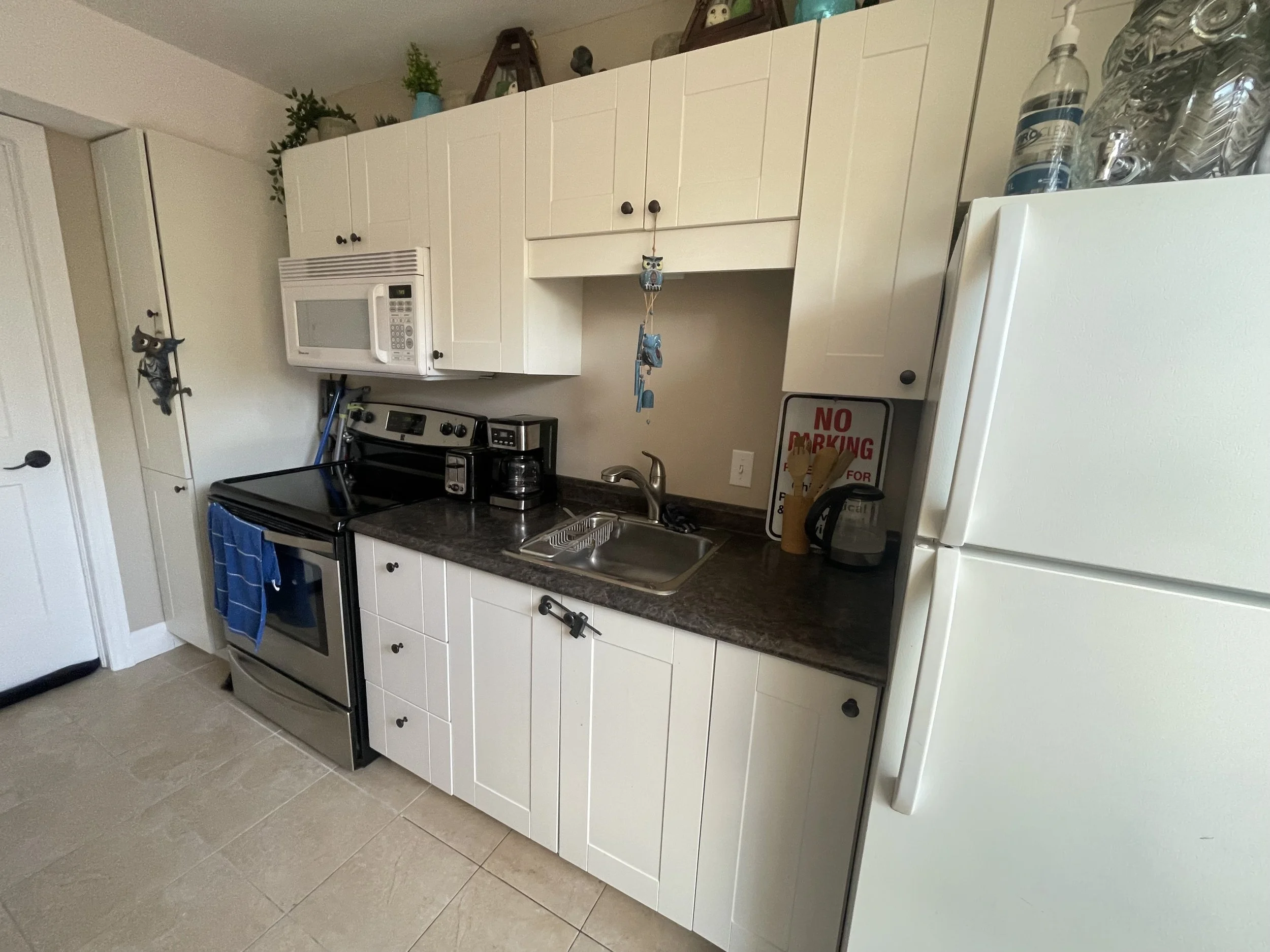Kitchen with white cabinets, stove, microwave, coffee maker, refrigerator, and a 'No Parking' sign on the counter.