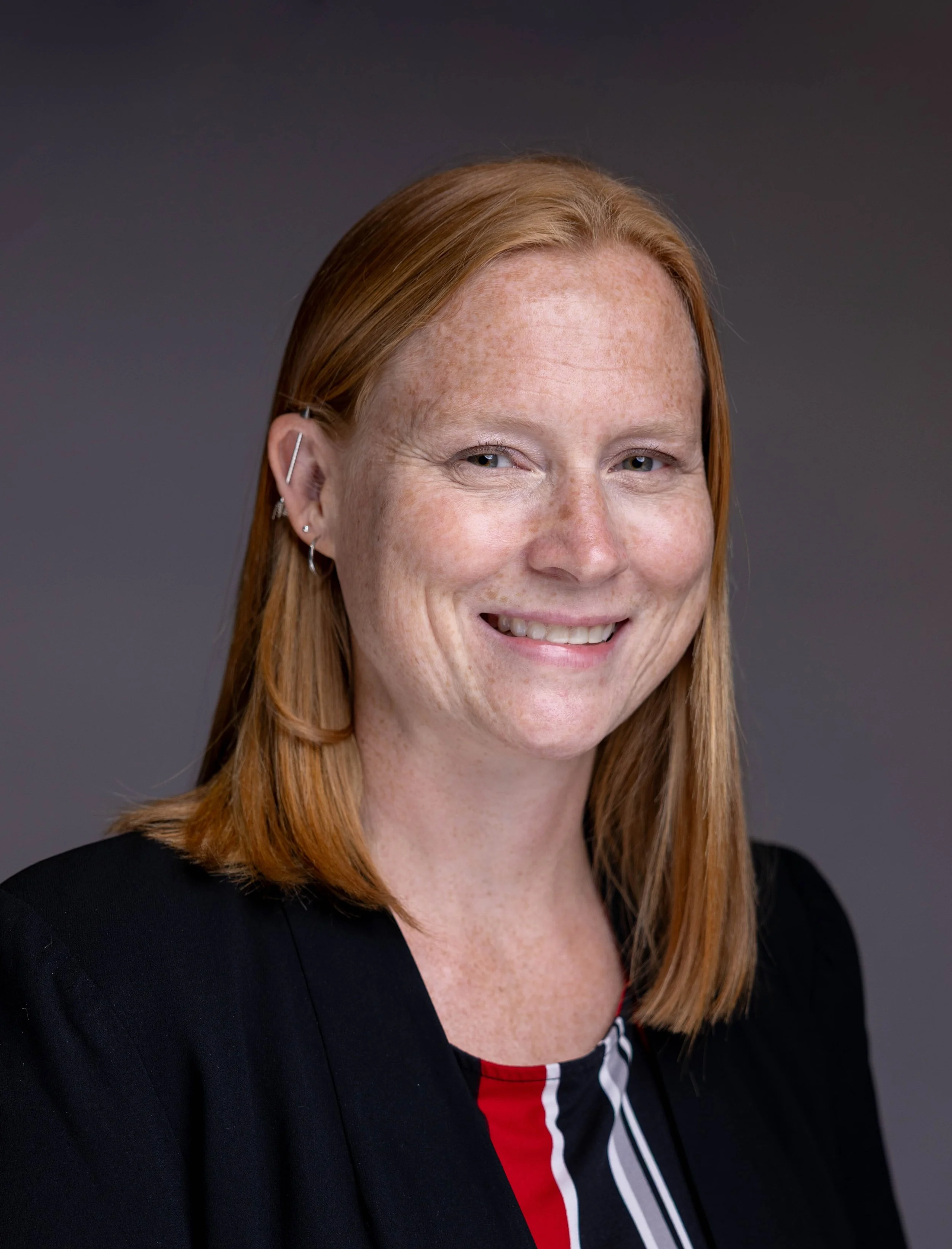A woman with red hair and freckles smiling at the camera, wearing earrings and a black blazer with a black, red, gray, and white patterned top, against a dark gray background.
