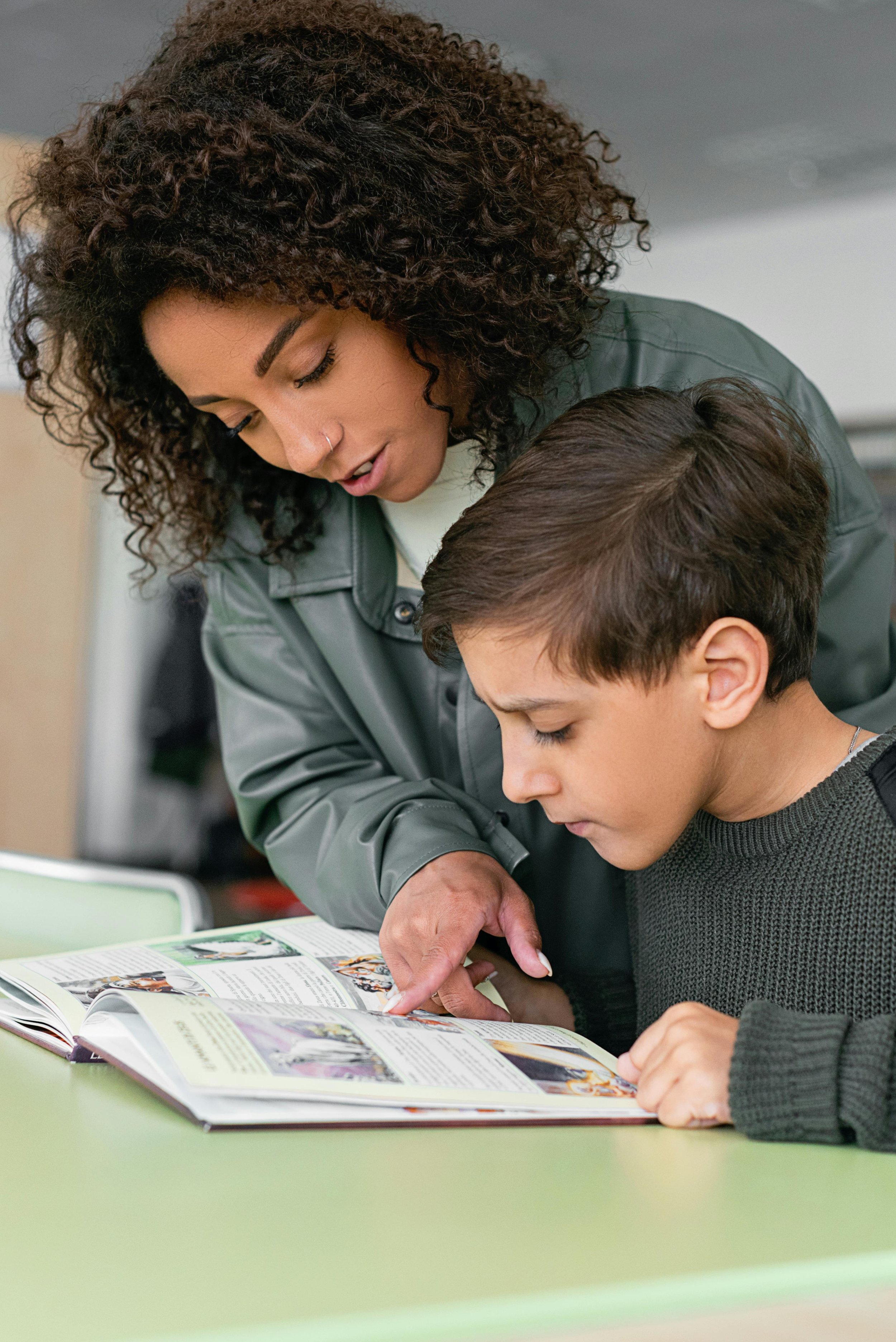A woman with curly hair and a nose piercing helping a young boy with short brown hair look at a colorful magazine or catalog on a green table.