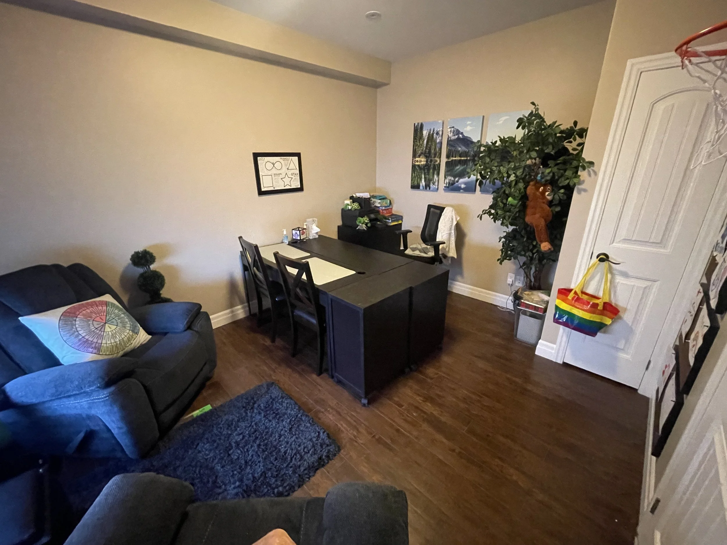A living room with a black sofa, a colorful cushion, a black dining table with chairs, a large potted plant, and wall art of a mountain and lake scene.