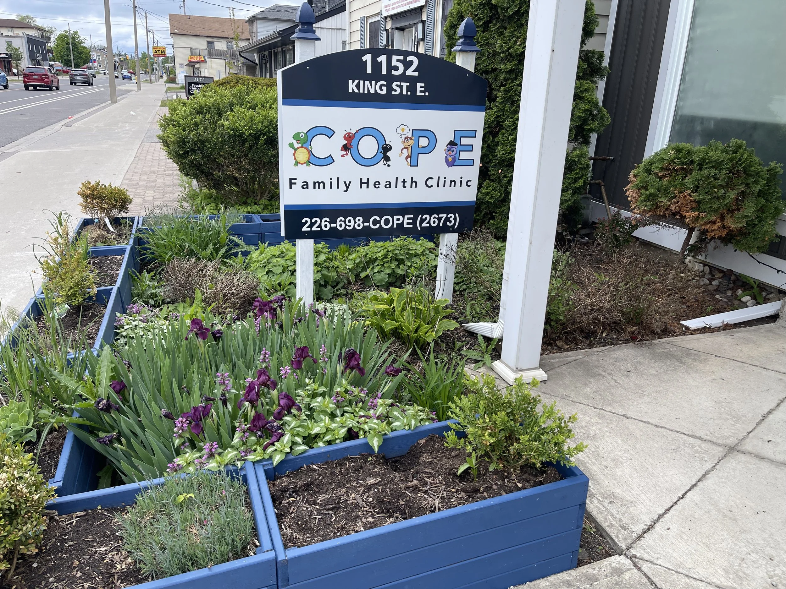 Sign for COPE Family Health Clinic in front of a garden with purple flowers and green plants, located at 1152 King St. E., with a phone number 226-698-COPE.