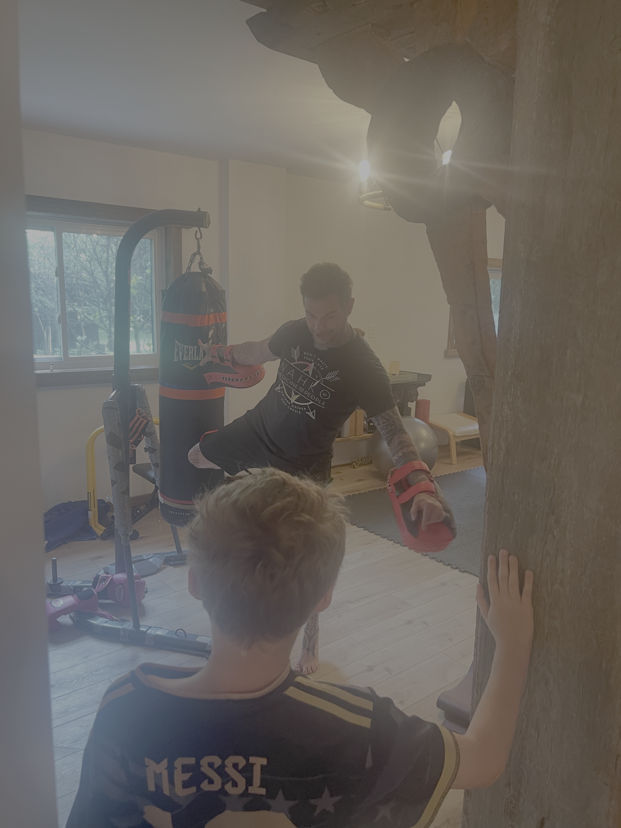A man practicing boxing with focus mitts while a young boy touches a large tree trunk in a home gym with exercise equipment.