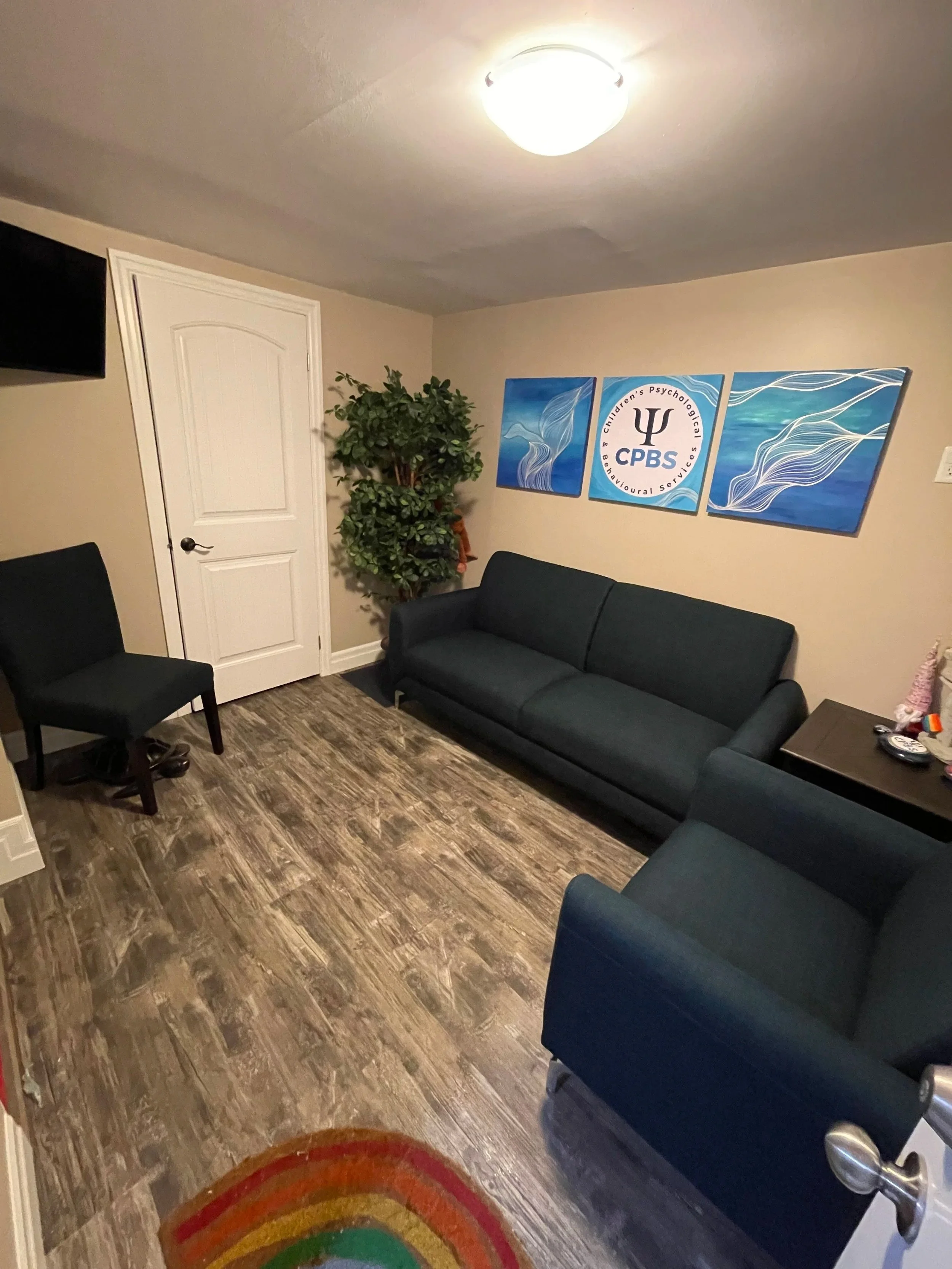 A living room with a black sofa, a black armchair, a small table with objects, a potted plant, a three-panel wall art depicting blue and white ocean waves with the CPBS logo in the center, a door, a wall-mounted TV, and a colorful rainbow rug on wood-look flooring.