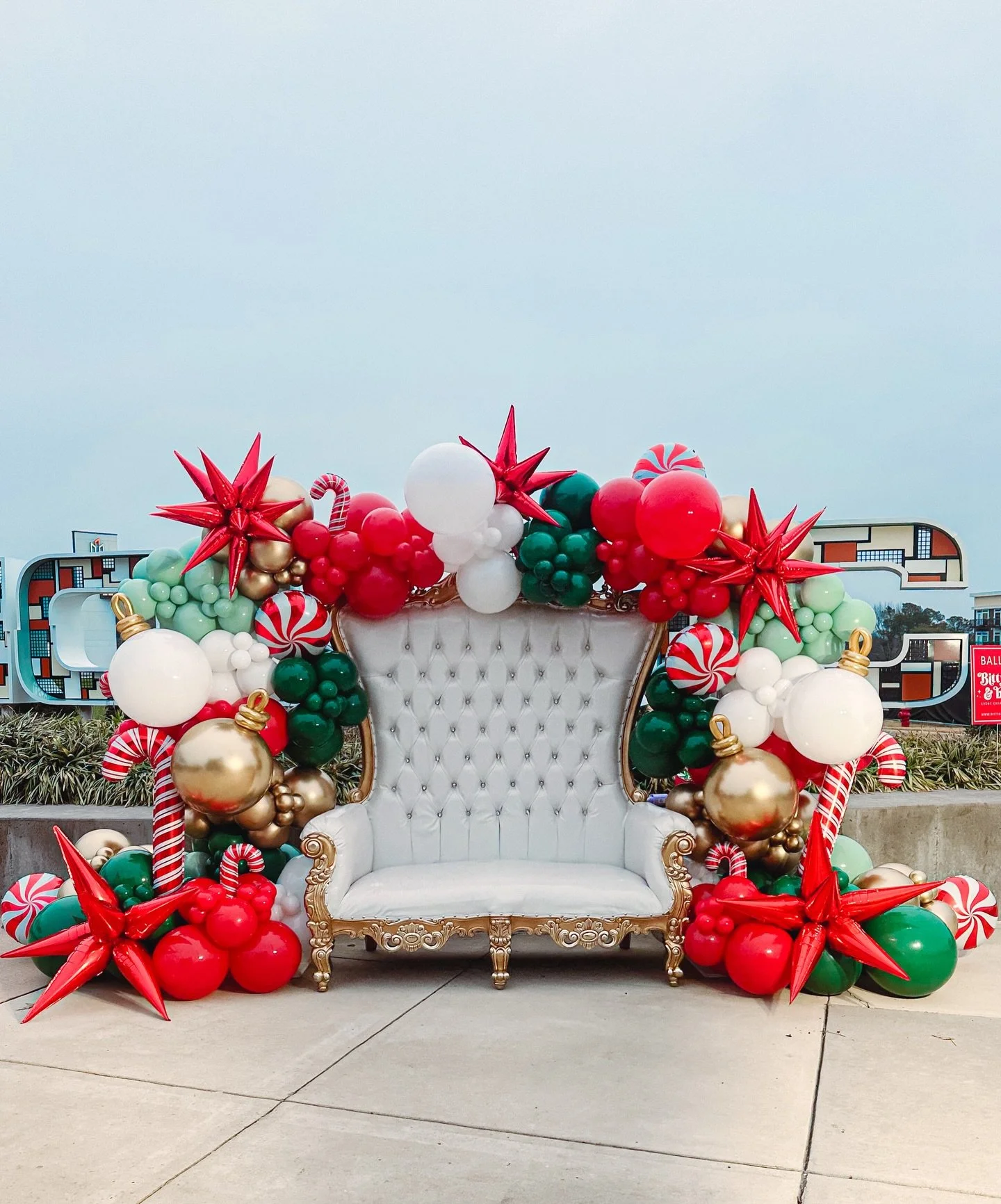 White vintage sofa decorated with Christmas balloons in red, green, white, gold, and candy cane patterns, arranged as a holiday display outdoors.