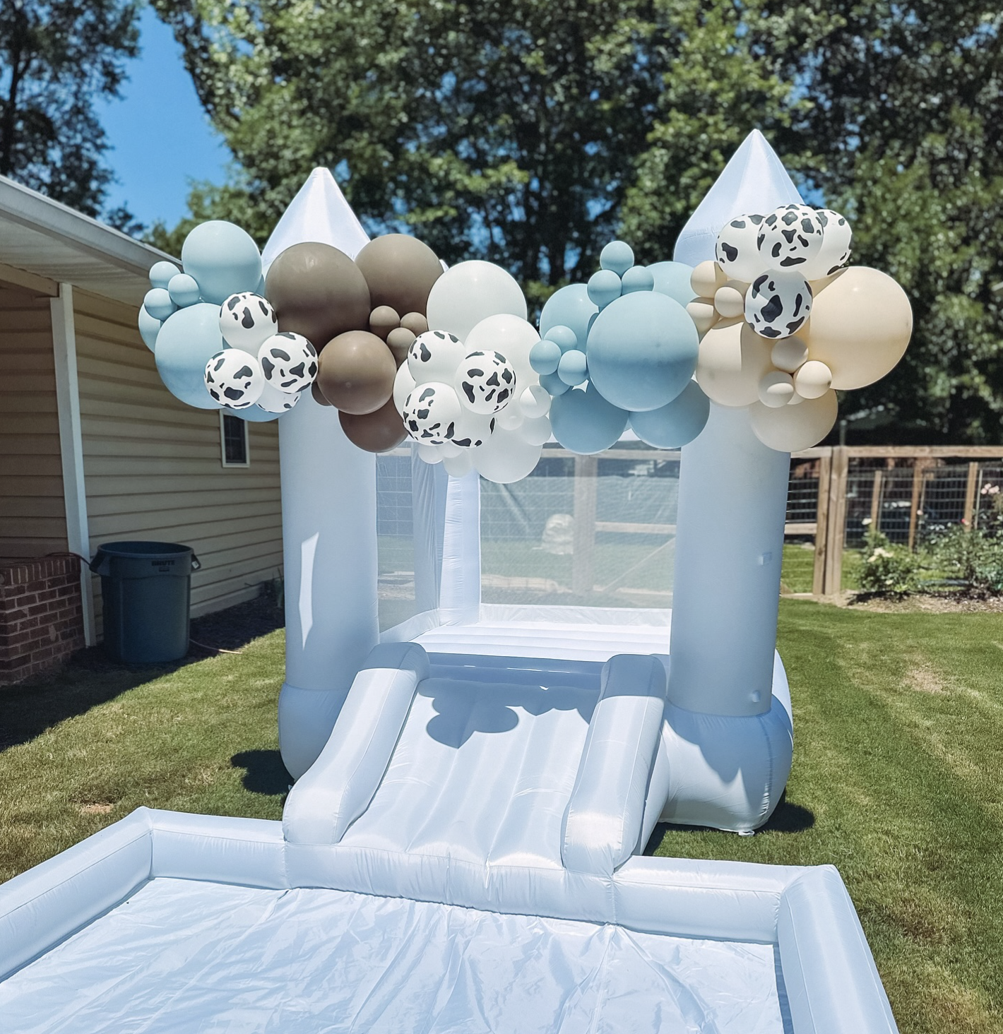 Inflatable white bounce house with blue, beige, and patterned balloons attached to the top, set up outdoors on grass near a house with trees in the background.
