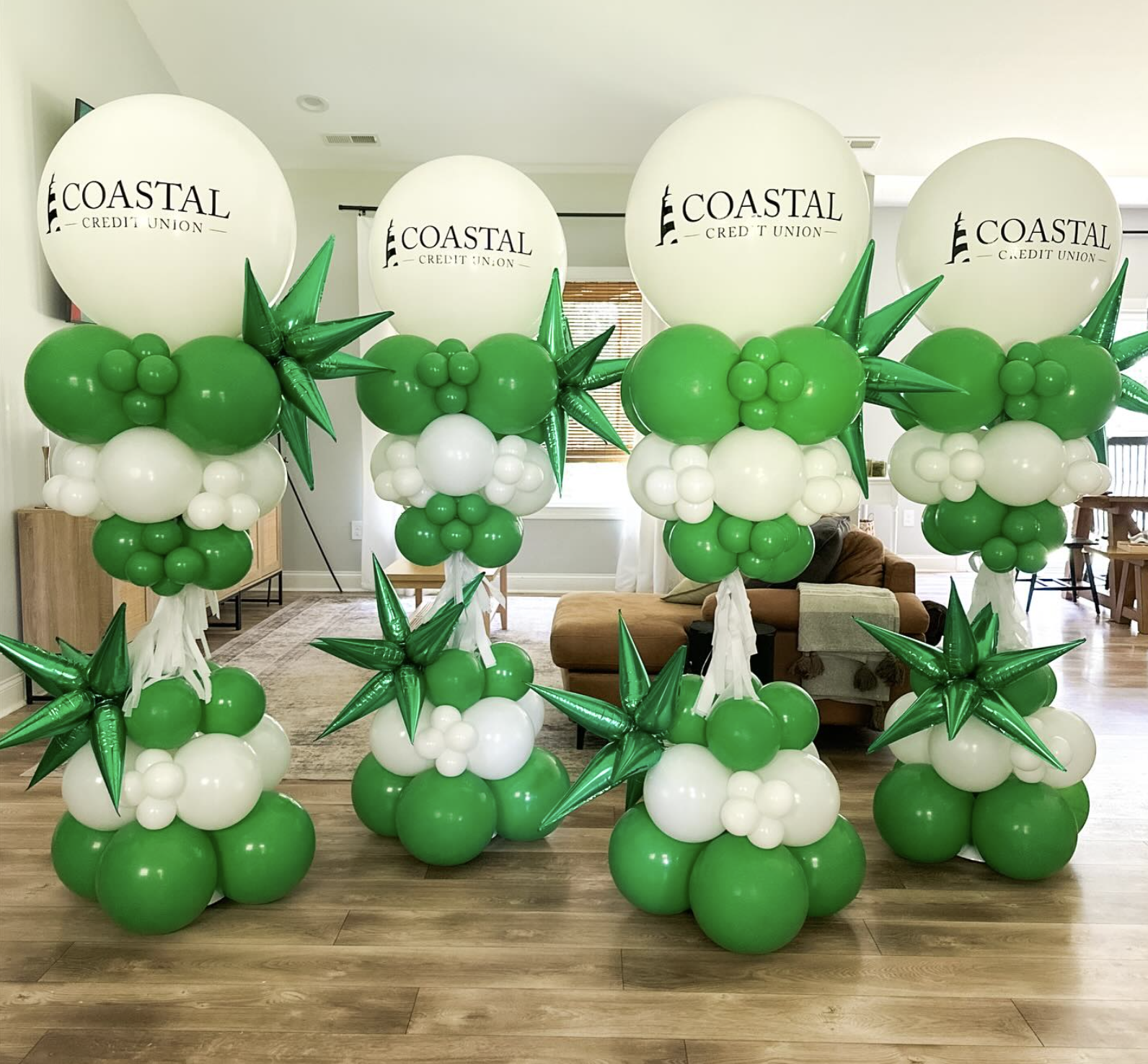 Group of green and white balloon arrangements with large white balloons featuring the Coastal Credit Union logo, decorated with green star-shaped and round balloons, placed in front of a living room with wooden floors and furniture.