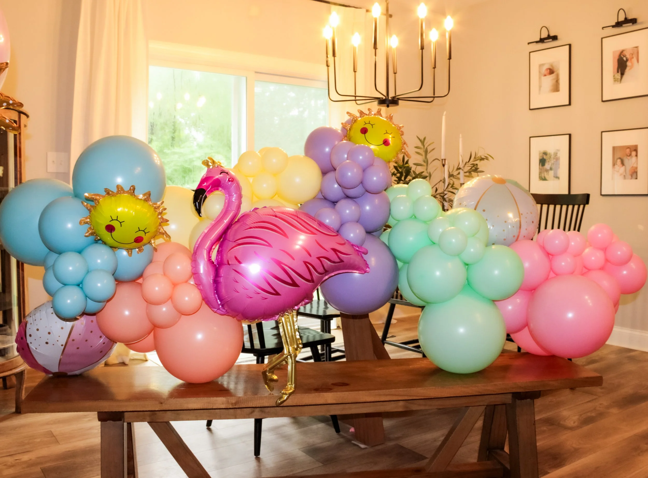 Colorful balloons and decorations on a wooden table inside a room with a chandelier, wall pictures, and large windows.