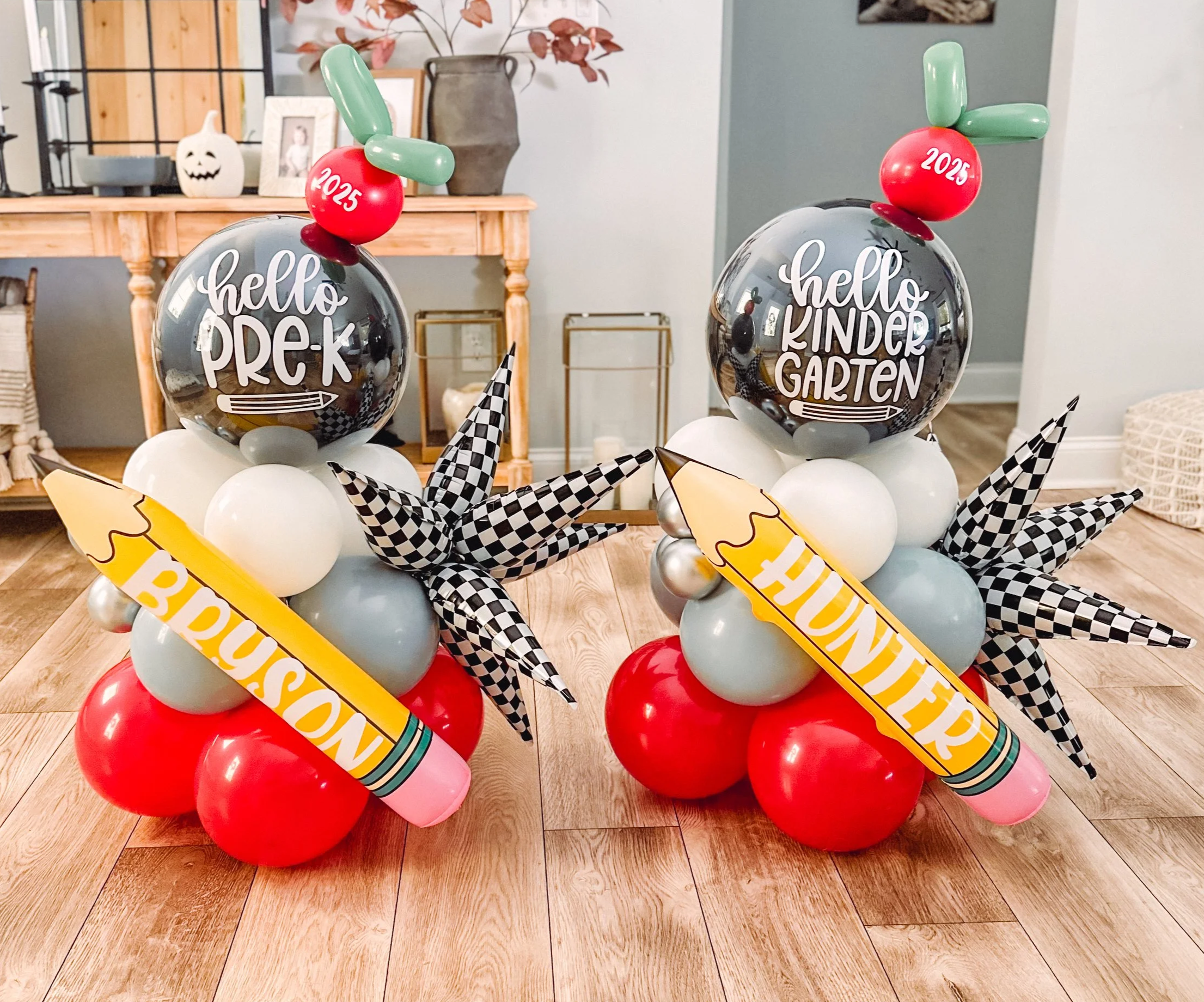 Two balloon arrangements for a preschool event, each with a large clear balloon with white text, smaller red, white, and gray balloons, checkered star-shaped balloons, and large yellow pencil-shaped balloons with white text. One says 'hello PRE-K' an